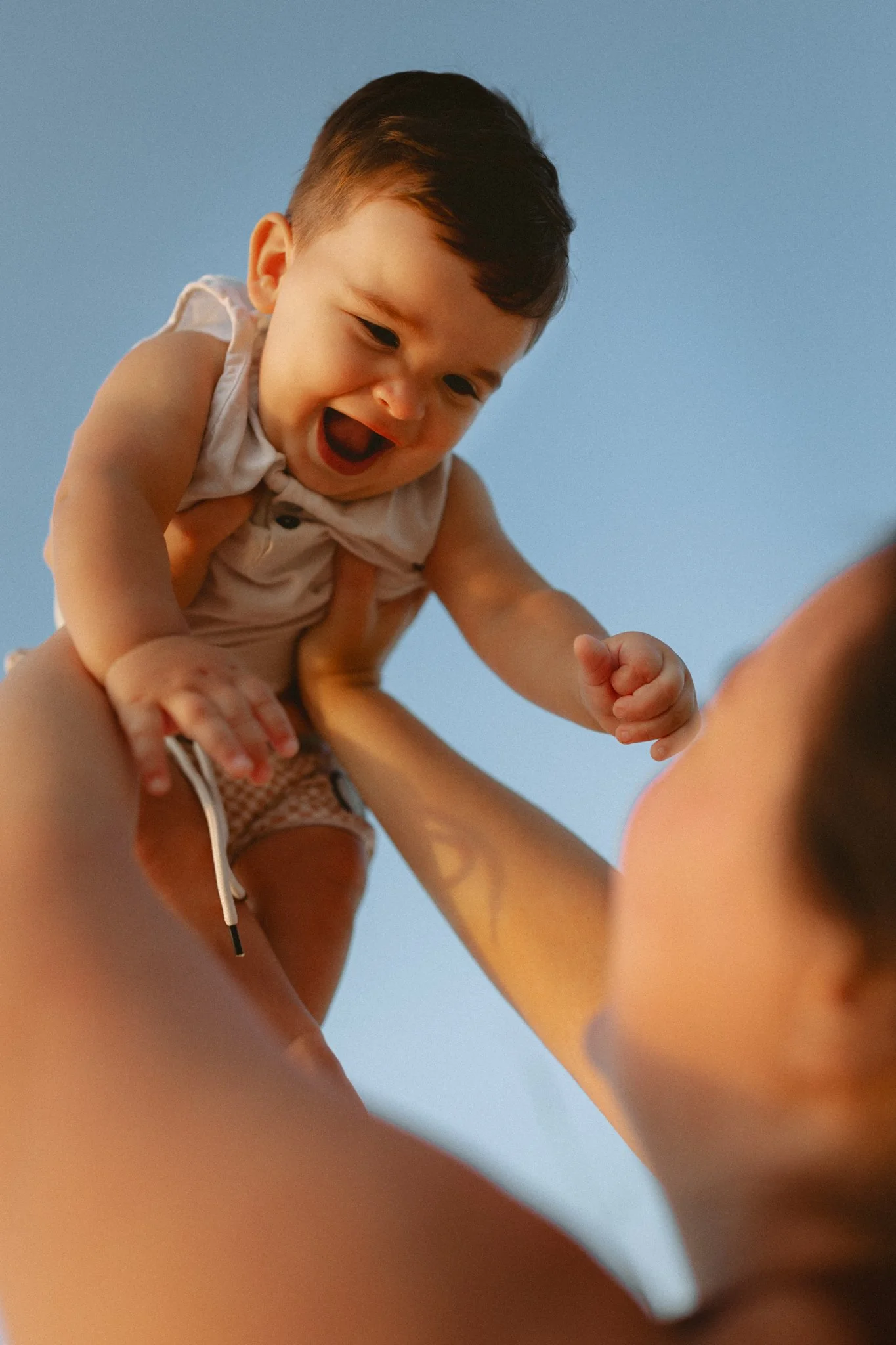 A young child with dark hair being lifted into the air, smiling, with an adult partially visible against a clear blue sky.