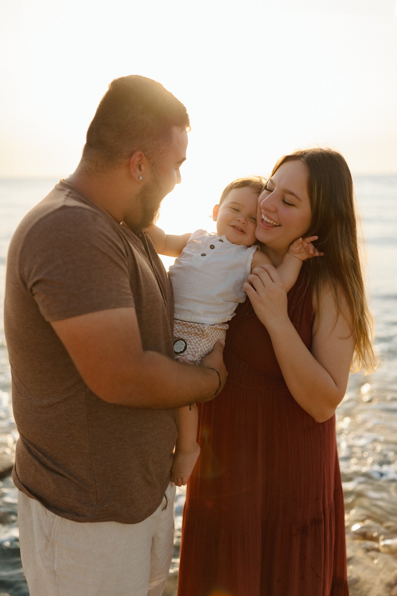 A happy family of three at the beach during sunset, with the mother and father holding their young child and all three smiling.