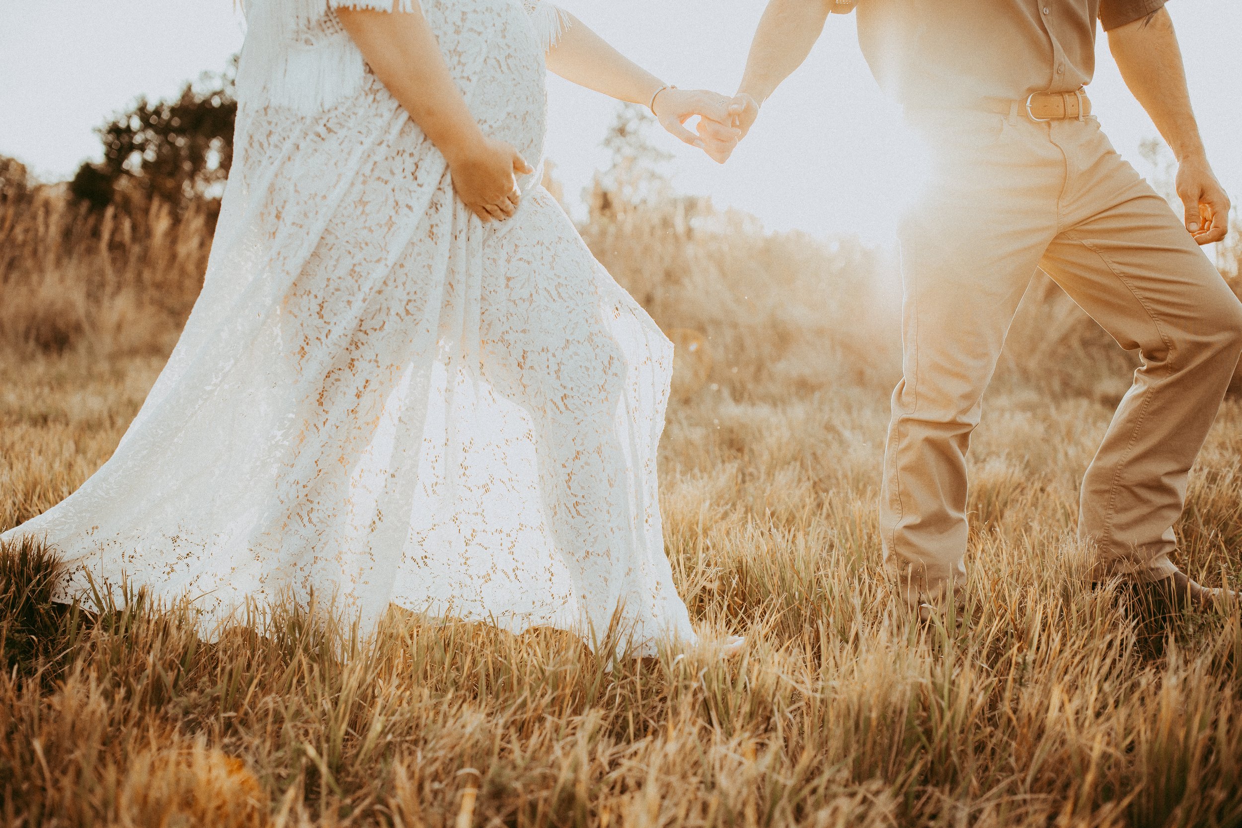 A couple holding hands in a field at sunset, with the woman wearing a white lace dress and the man in tan pants and a shirt.