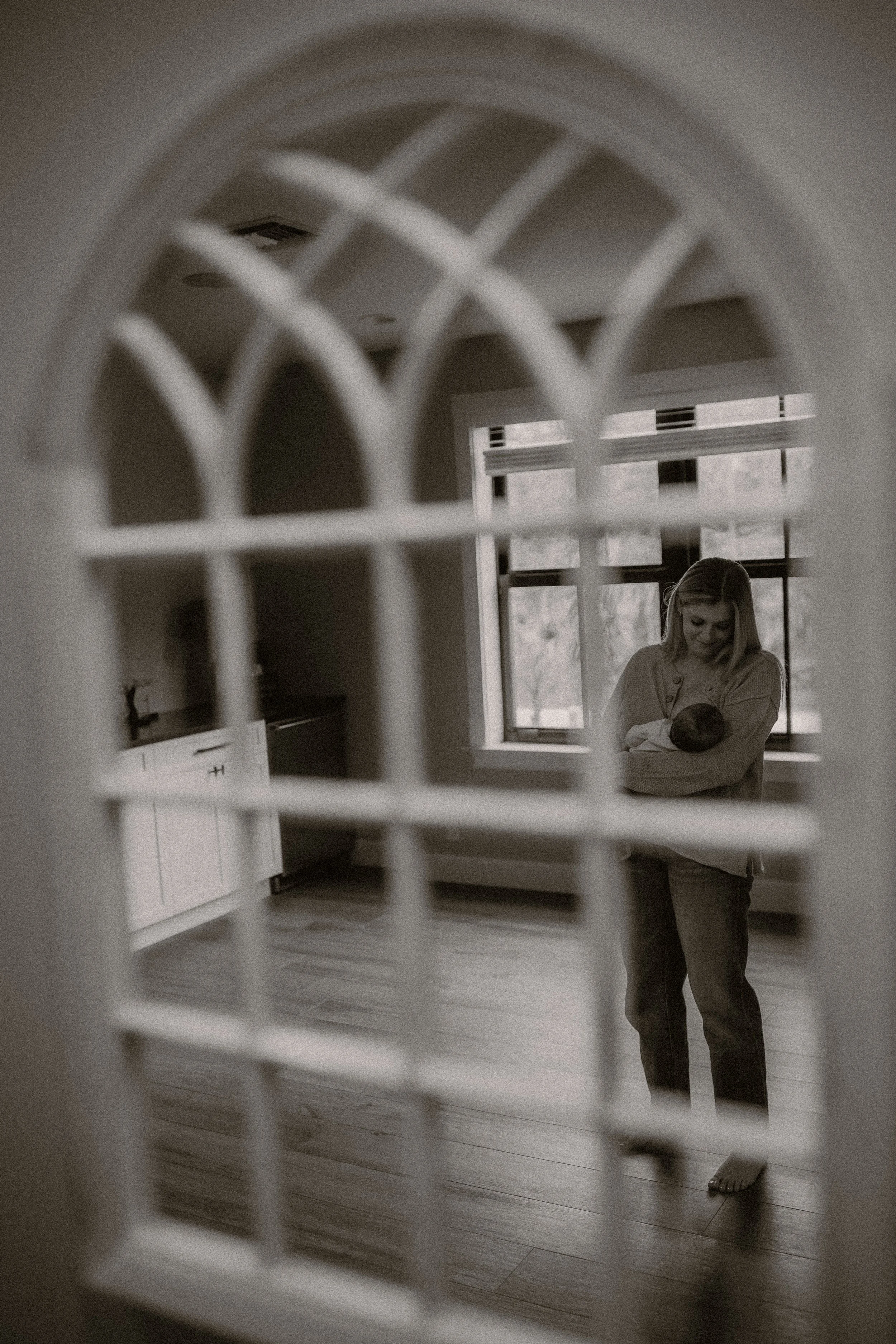 A woman smiling and holding a newborn baby, seen through a decorative window with a grid pattern, in a room with large windows and natural light.