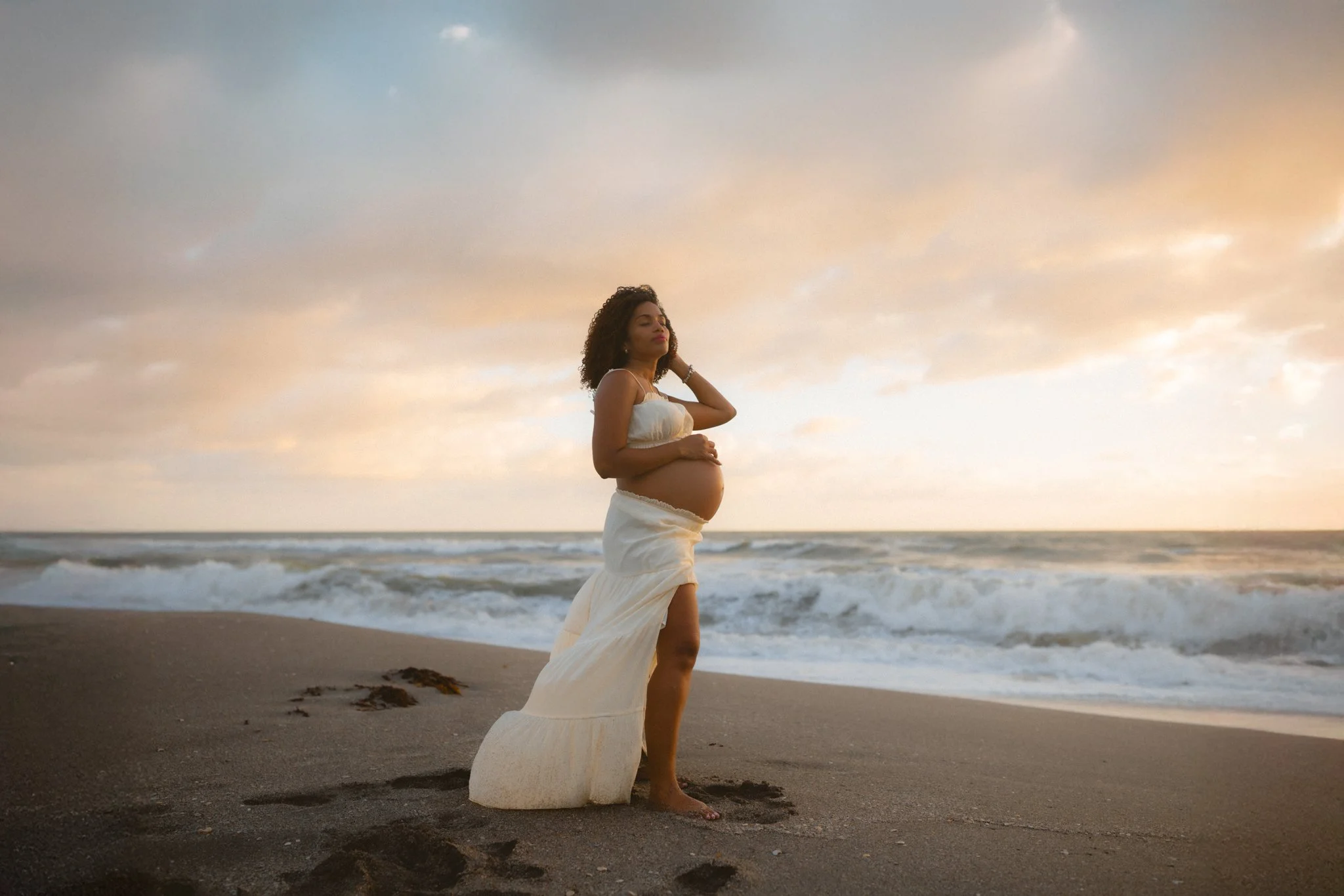 Pregnant woman standing on the beach at sunset, wearing a white skirt and top, with her eyes closed and one hand on her stomach.