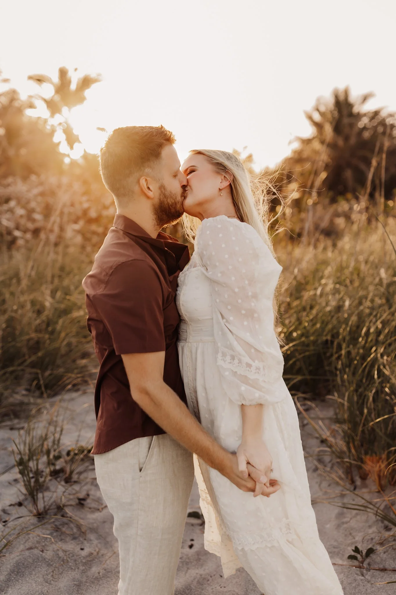 A couple kissing and holding hands on a beach at sunset, with palm trees and tall grass in the background.