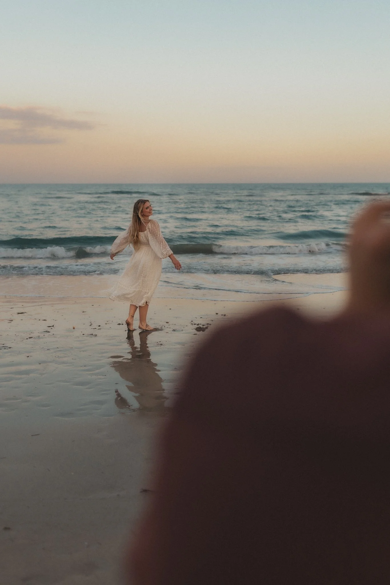 A woman in a white dress walking along the beach at sunset with the ocean in the background.
