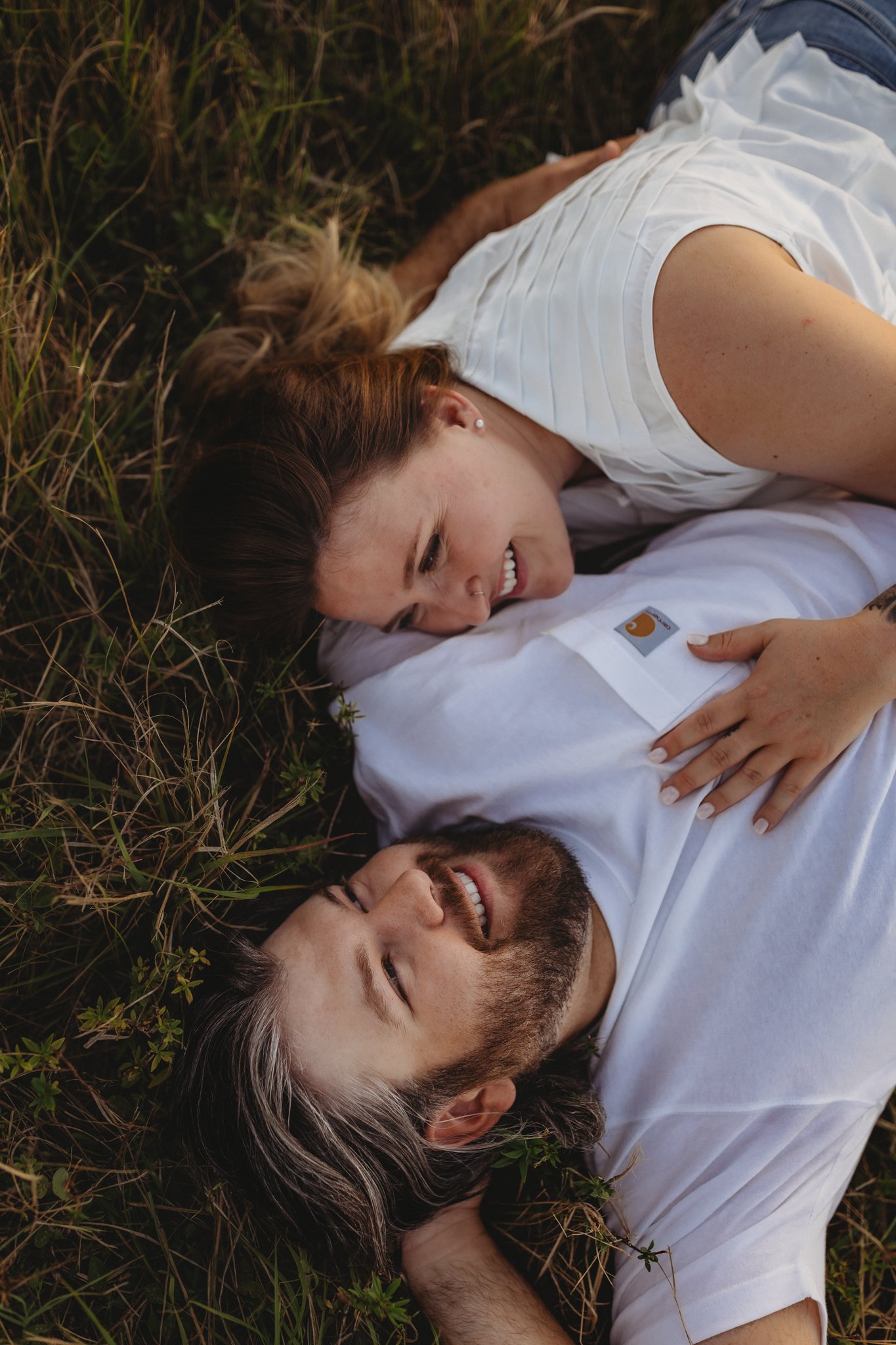 A couple lying on the grass, looking at each other and smiling, with the woman resting her hand on the man's chest.