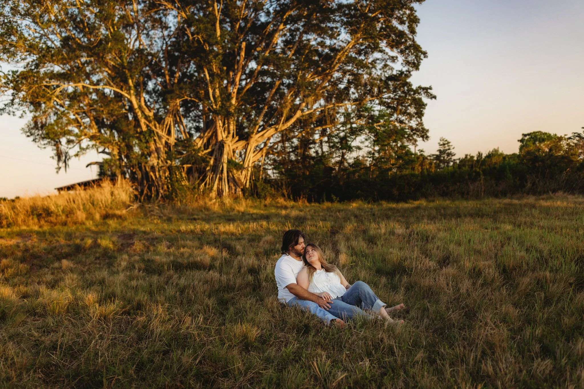 A couple sitting on grass in a field at sunset, with a large tree in the background.