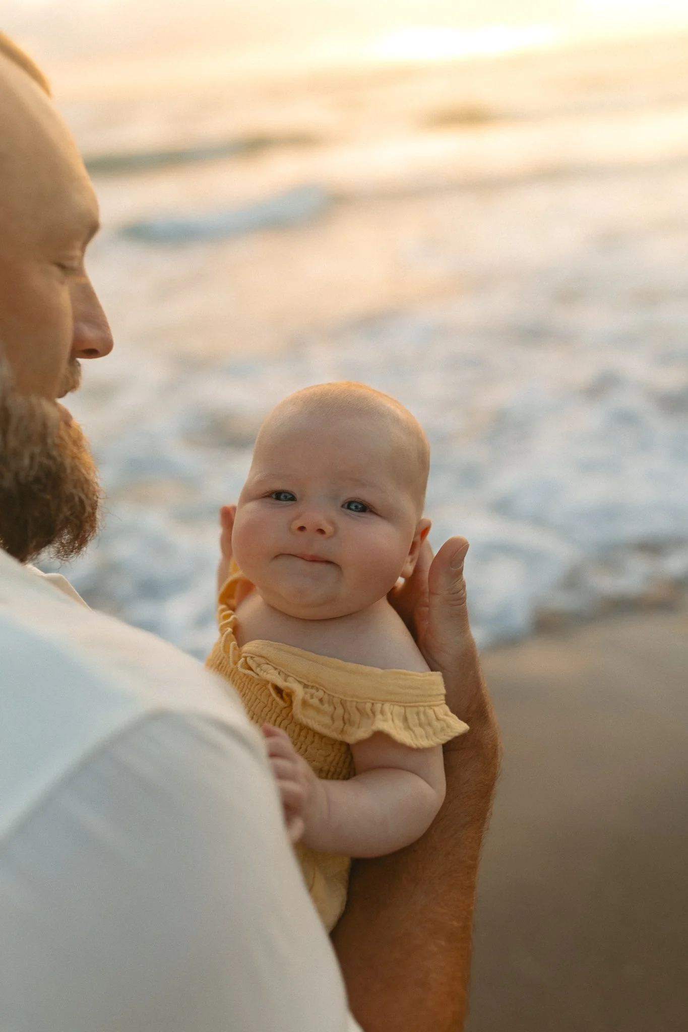 a father holdings his newborn baby wearing a  yellow outfit in front of the ocean 
