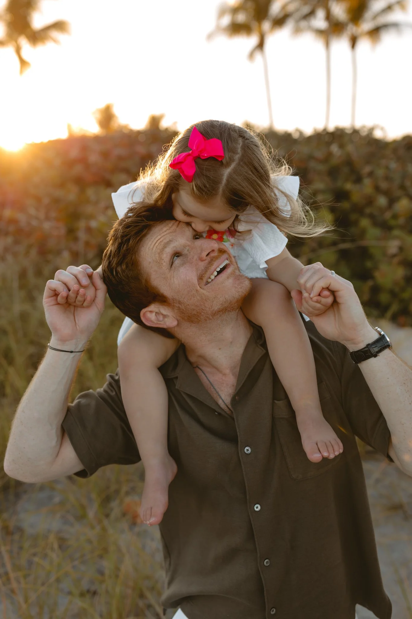 A man with red hair and a woman with a young girl riding on his shoulders outdoors at sunset, with trees and palm trees in the background.