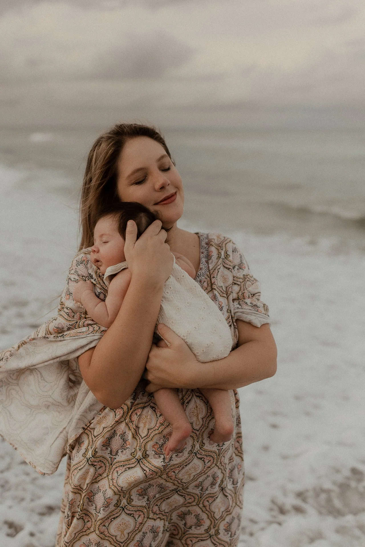 A woman holding a sleeping baby close to her chest on a beach with cloudy skies.