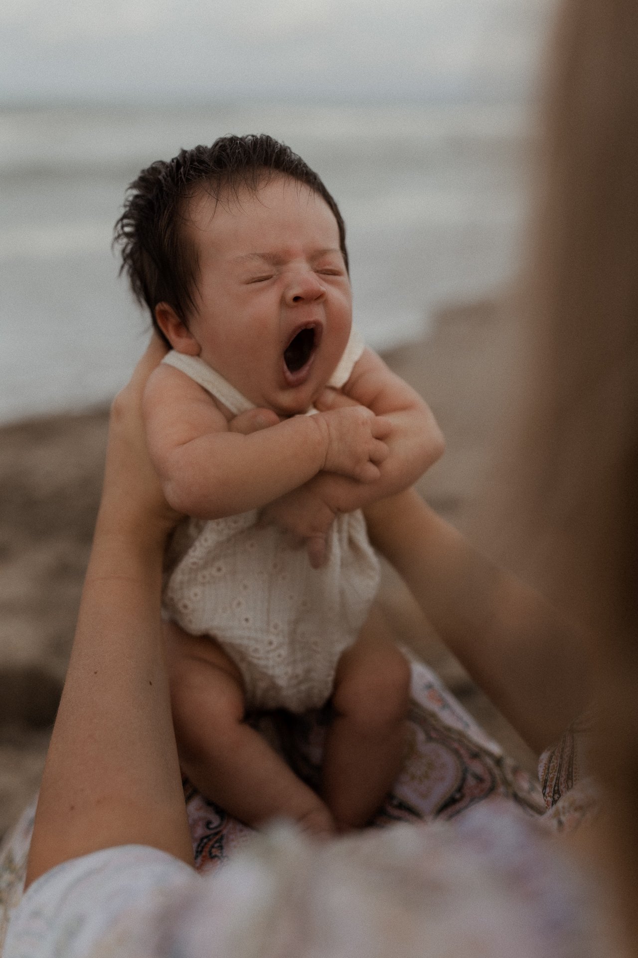 A baby yawning and stretching while being held by an adult on the beach with sand and water in the background.
