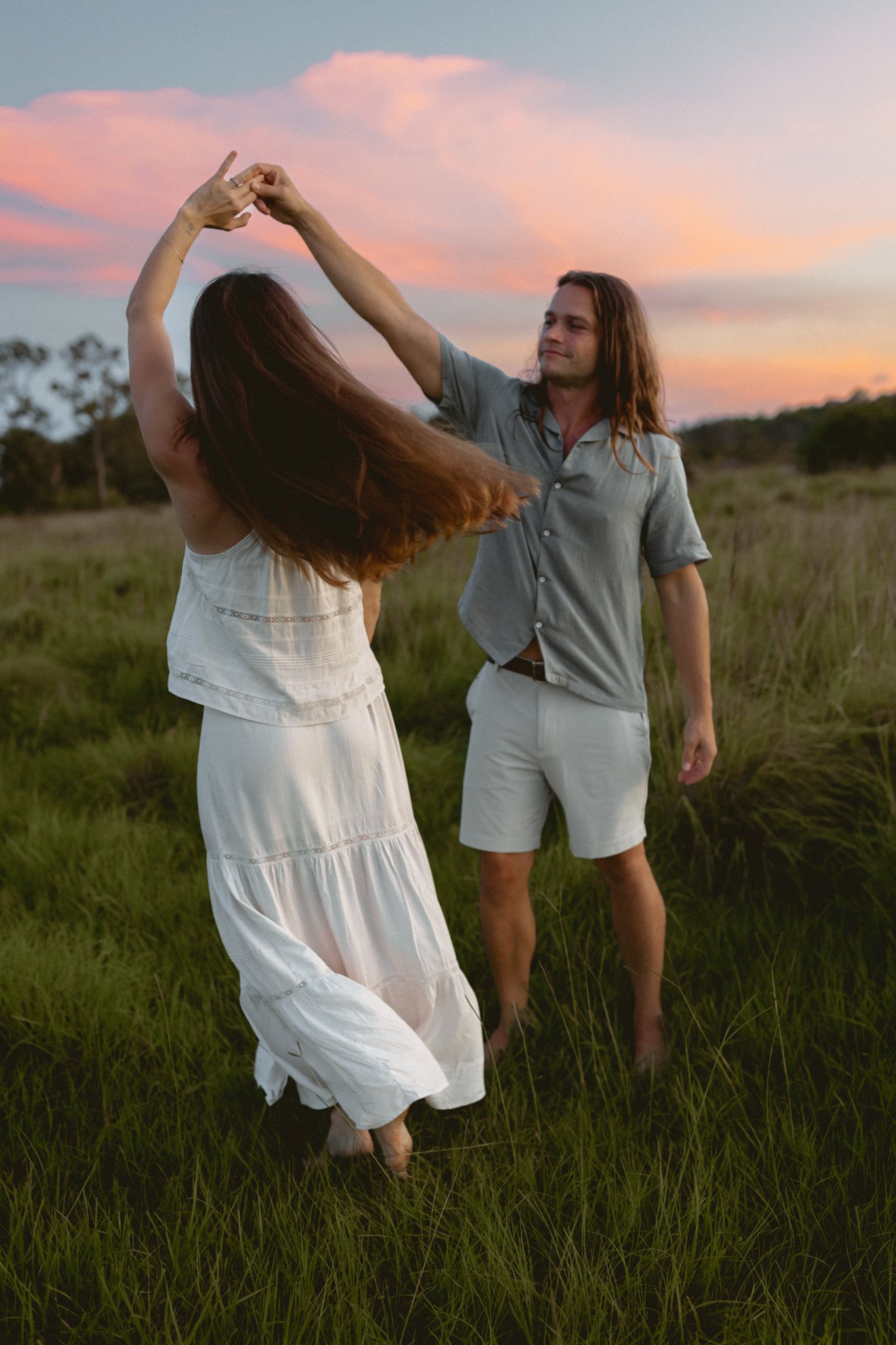 A young couple dancing barefoot in a grassy field during sunset, with pink and orange skies in the background.