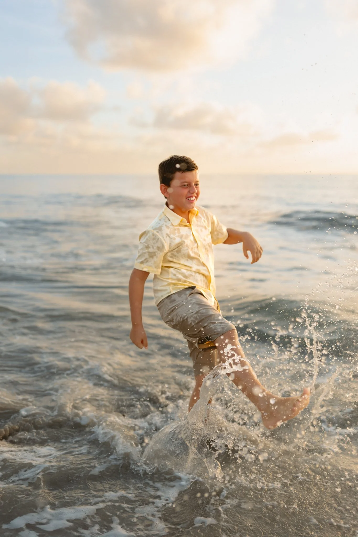 A young boy in a yellow shirt and khaki shorts playing in the ocean at sunset, splashing water and smiling.