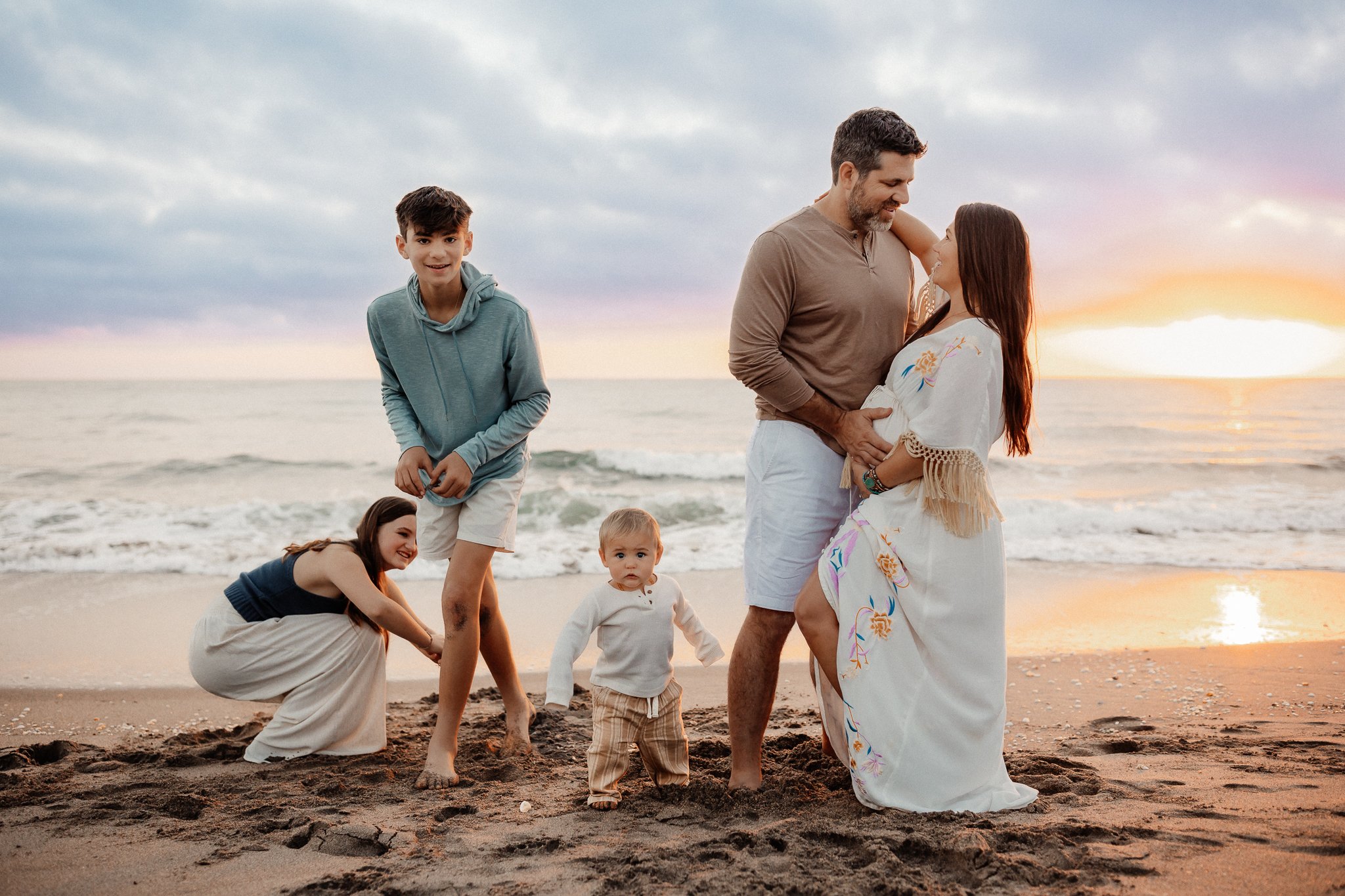 A family on the beach during sunset. A man and woman are embracing, looking at each other. Three children are playing nearby: a girl squatting, a boy standing, and a toddler walking. The sky has clouds with hues of pink, purple, and orange.