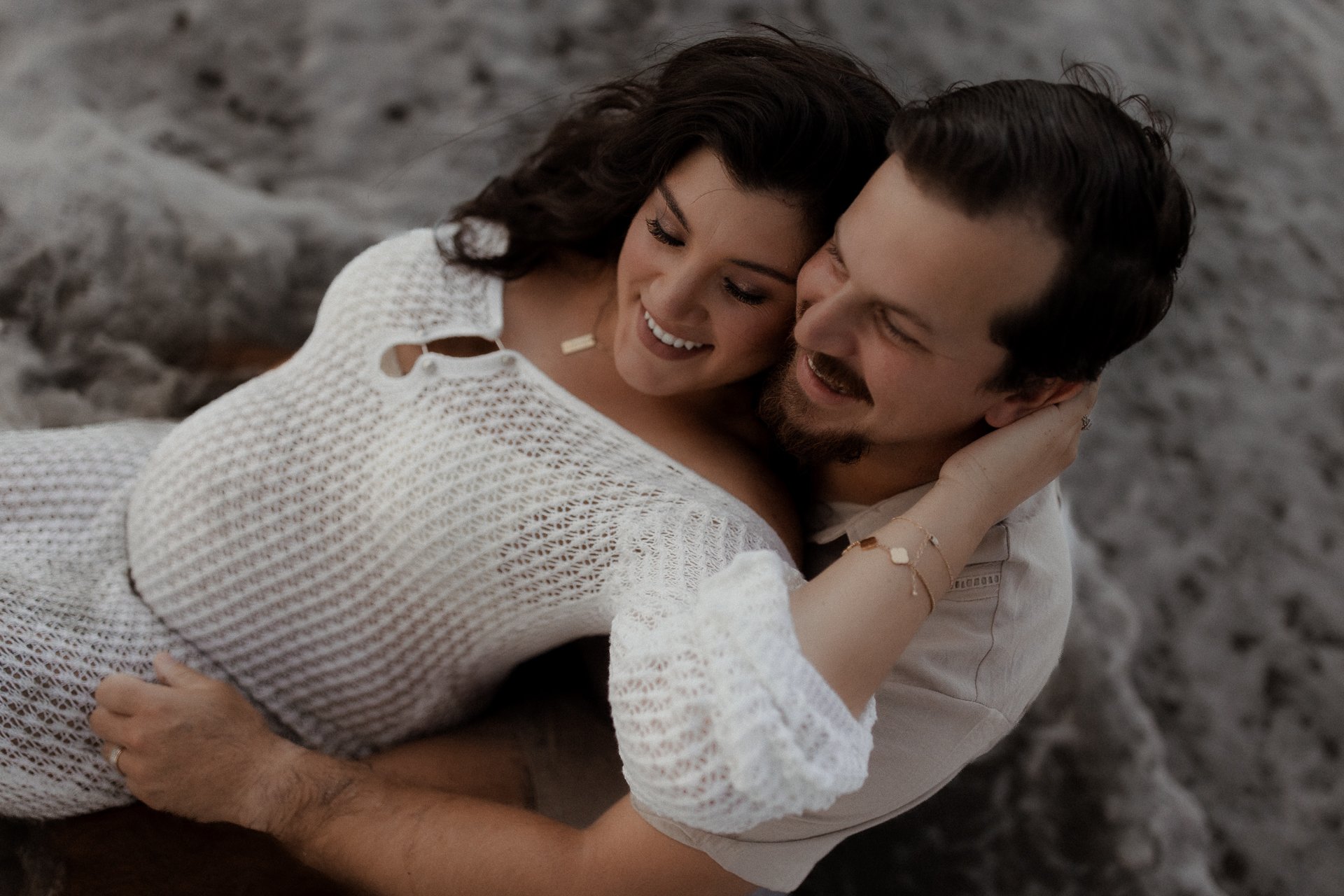 A smiling woman and man are cuddling, with the woman resting her head on the man's shoulder. They are lying on a rocky surface outdoors, embracing each other closely.