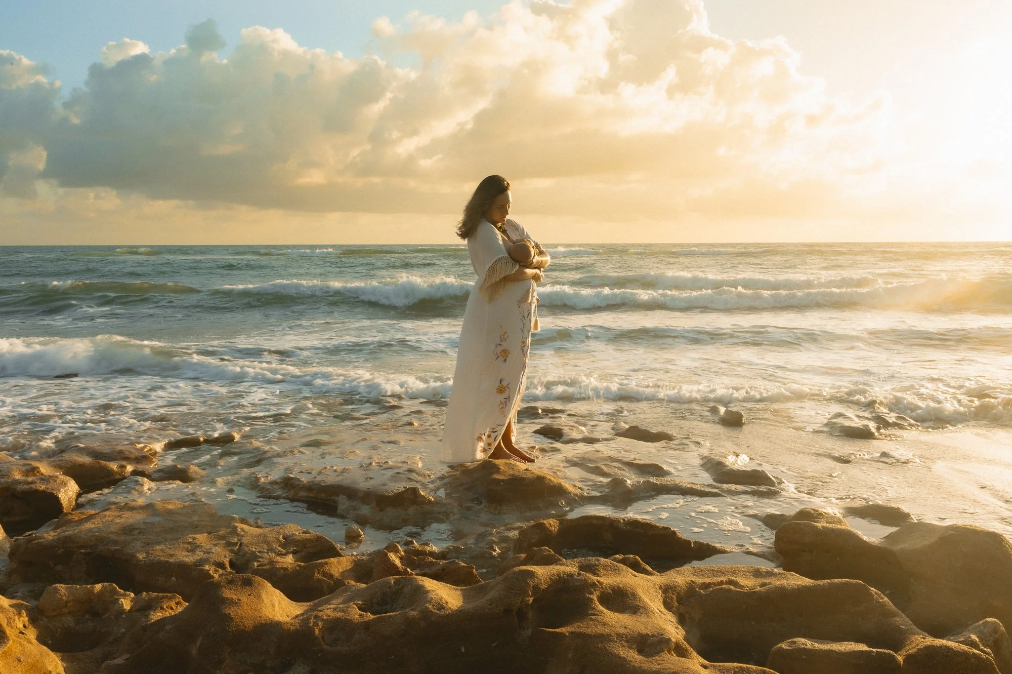 a mother breastfeeding her newborn daughter while standing on rocks at the beach at sunrise