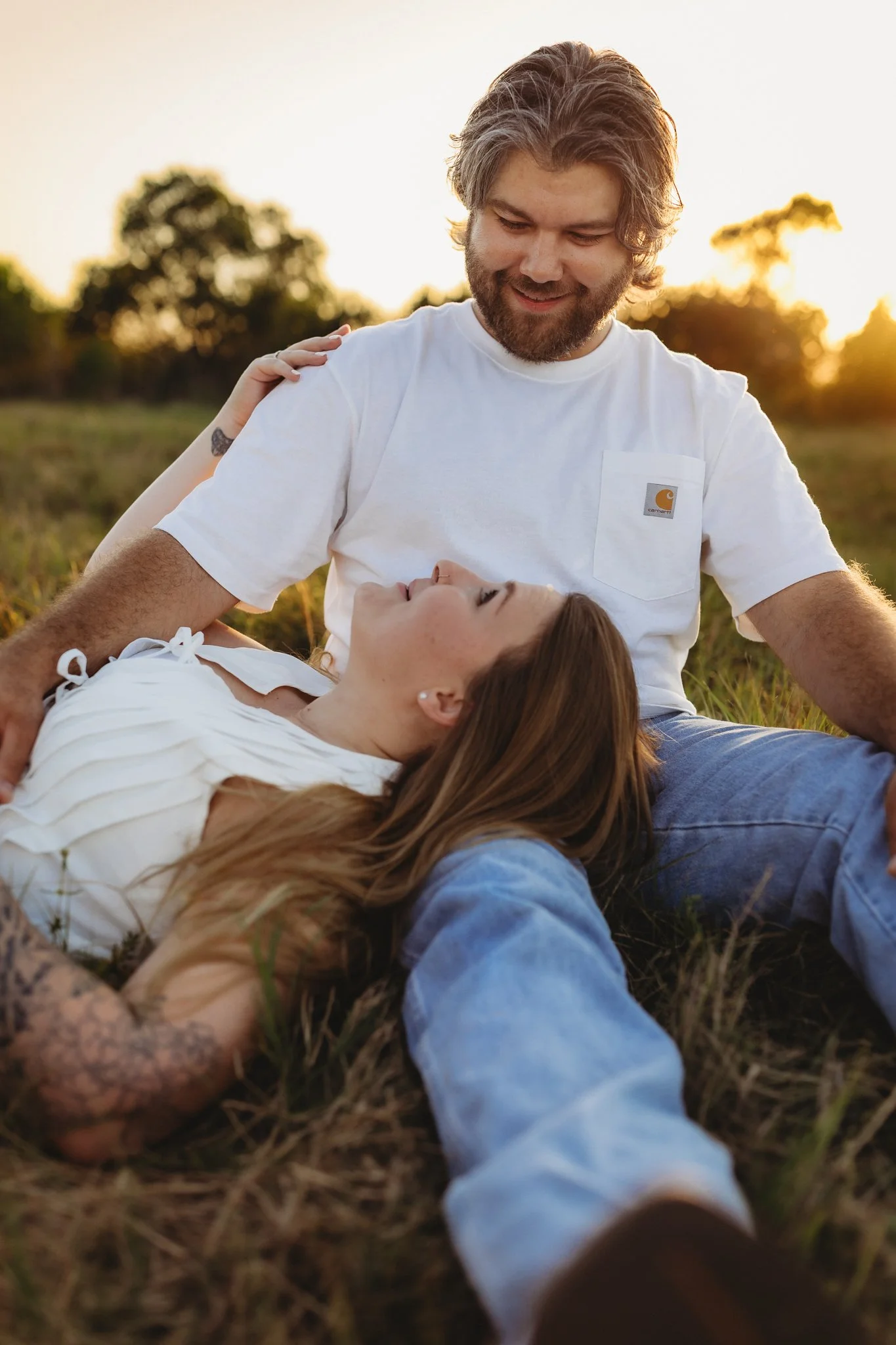 A man and woman sitting on grass outdoors at sunset, with the woman lying down and the man sitting next to her, smiling and looking down at her.