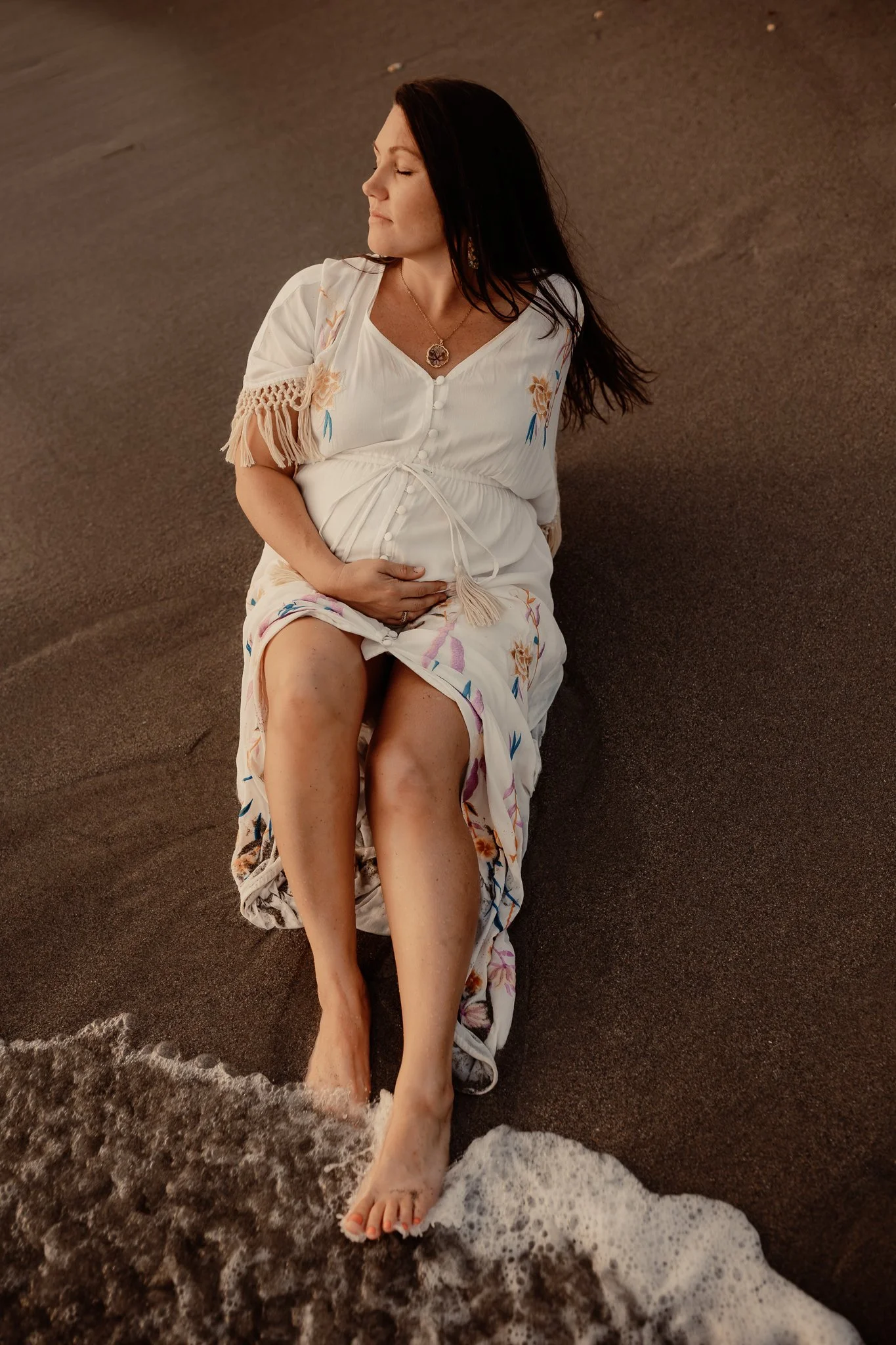 A woman sitting on the beach with her feet in the water, wearing a white dress with colorful embroidery, and holding her belly, possibly pregnant.