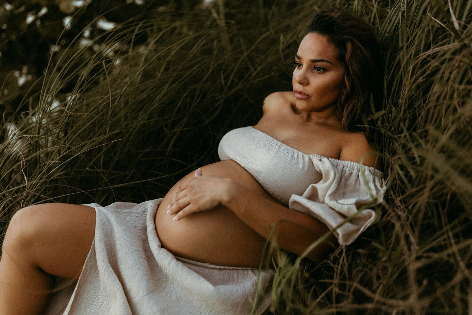 A pregnant woman lying outdoors on tall grass, wearing an off-shoulder light-colored dress, gently touching her belly and gazing thoughtfully to the side.