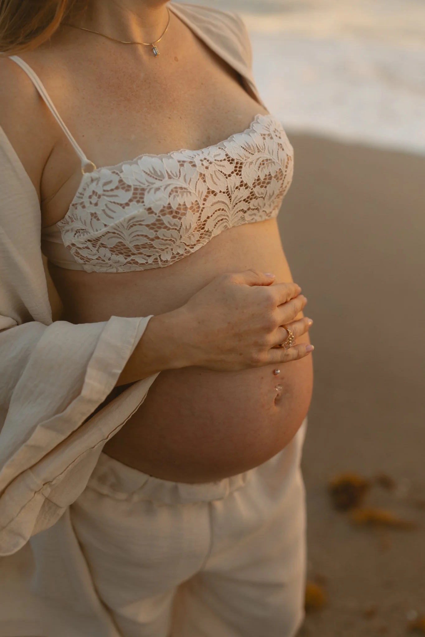 Pregnant woman holding her belly on the beach during sunset, wearing a white lace bralette and beige clothing.