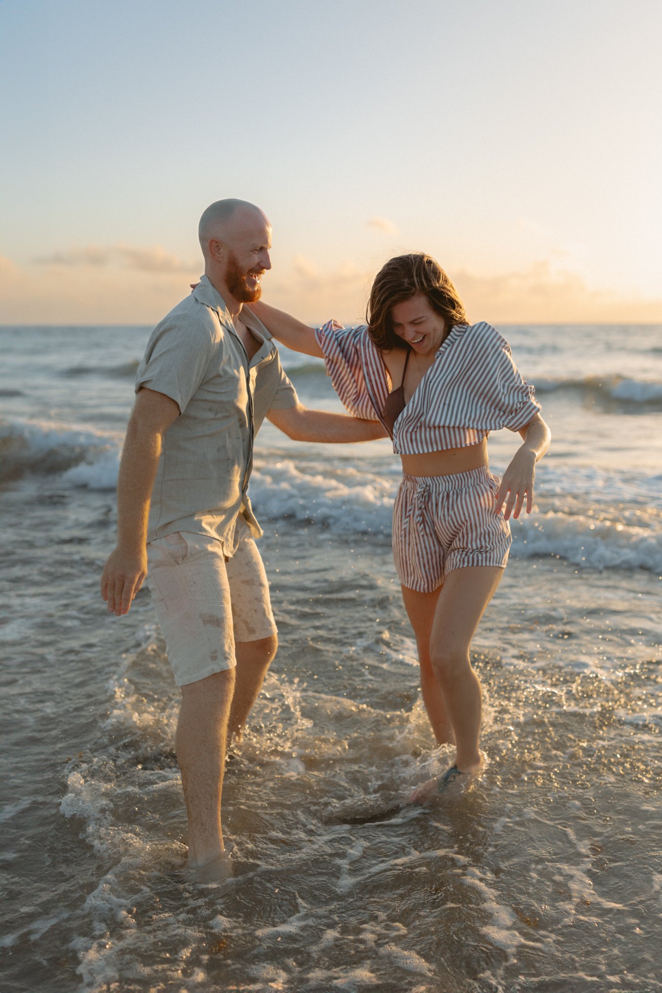 A couple enjoying a playful moment in the ocean at sunset, smiling and splashing water.