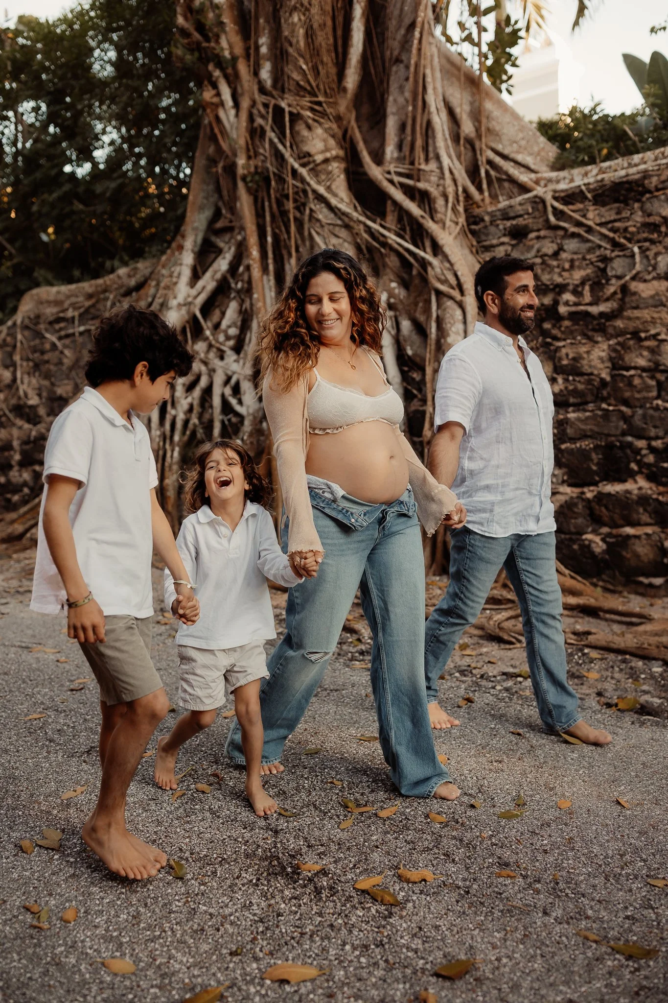 A pregnant woman and her family holding hands and walking barefoot outdoors near a large tree and stone wall, smiling and enjoying a sunny day.
