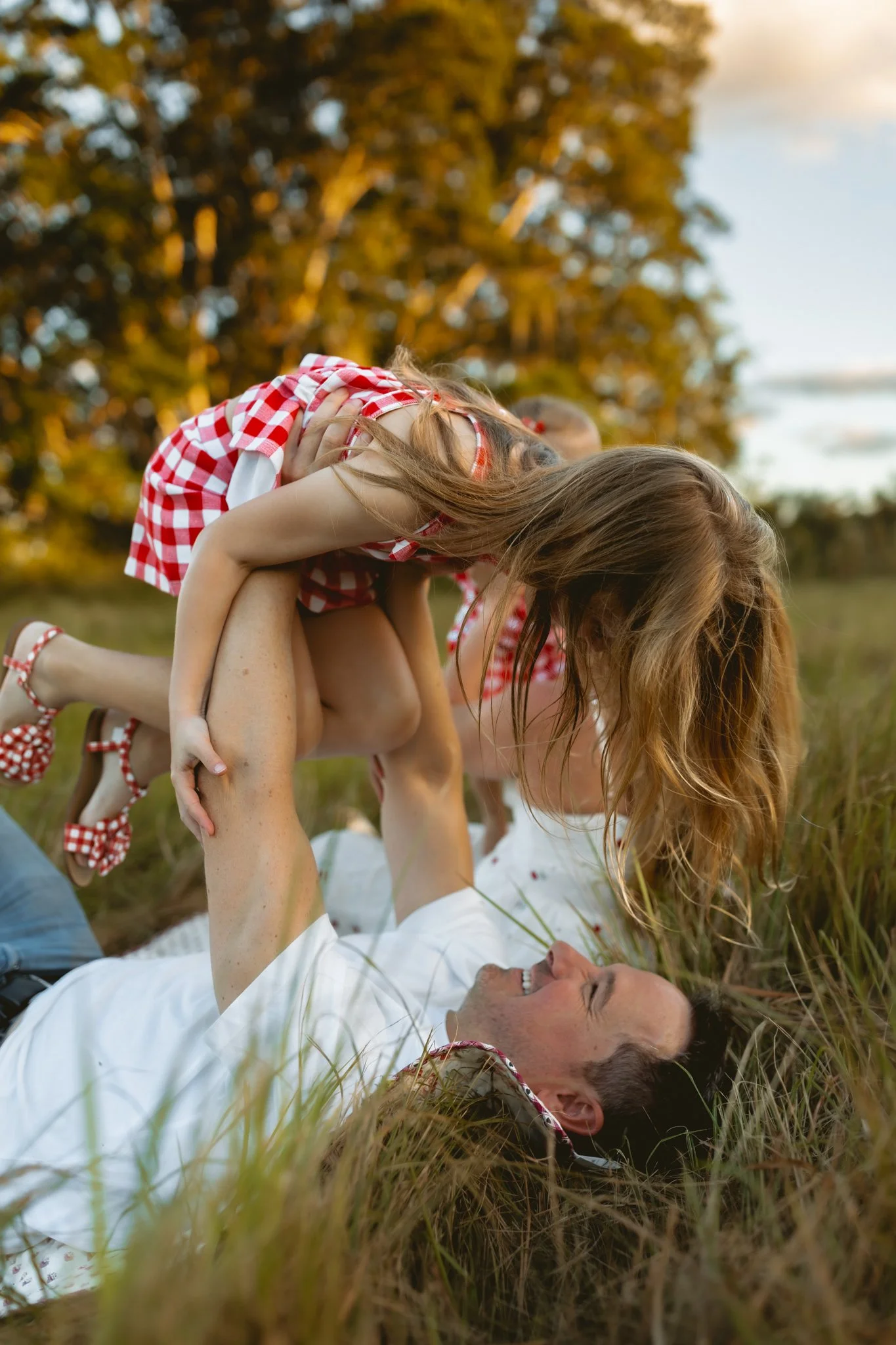 A man laying on the grass outdoors, smiling and playing with a young girl who is leaning over him. The girl is wearing a red and white checkered dress and polka dot shoes, and they are surrounded by trees and tall grass during sunset.