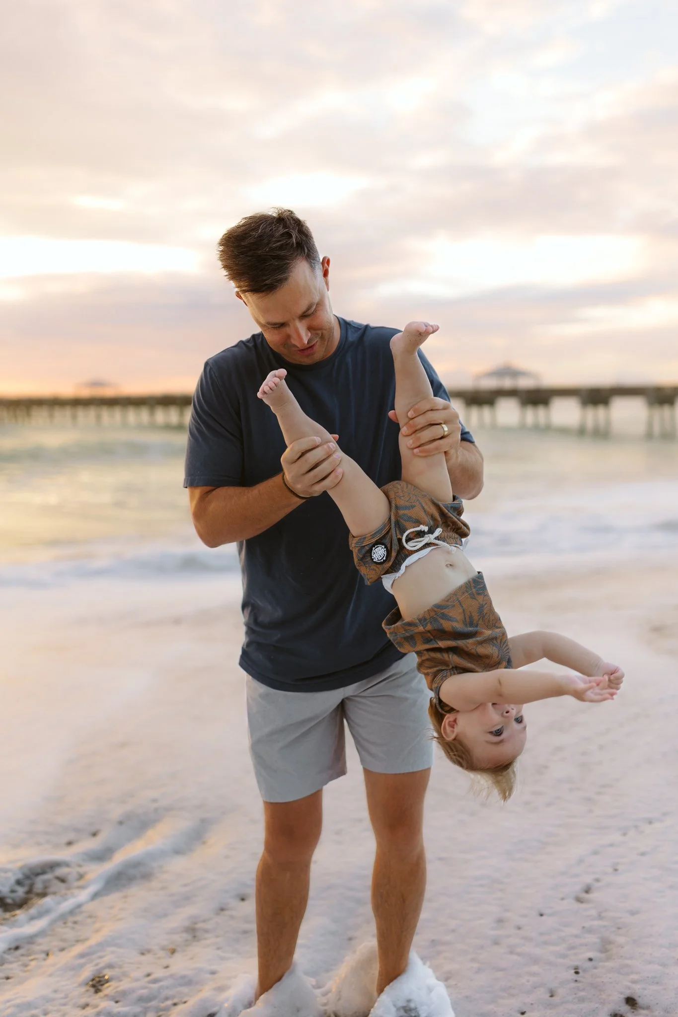 A man plays with a young child upside down on the beach during sunset, with a pier in the background.