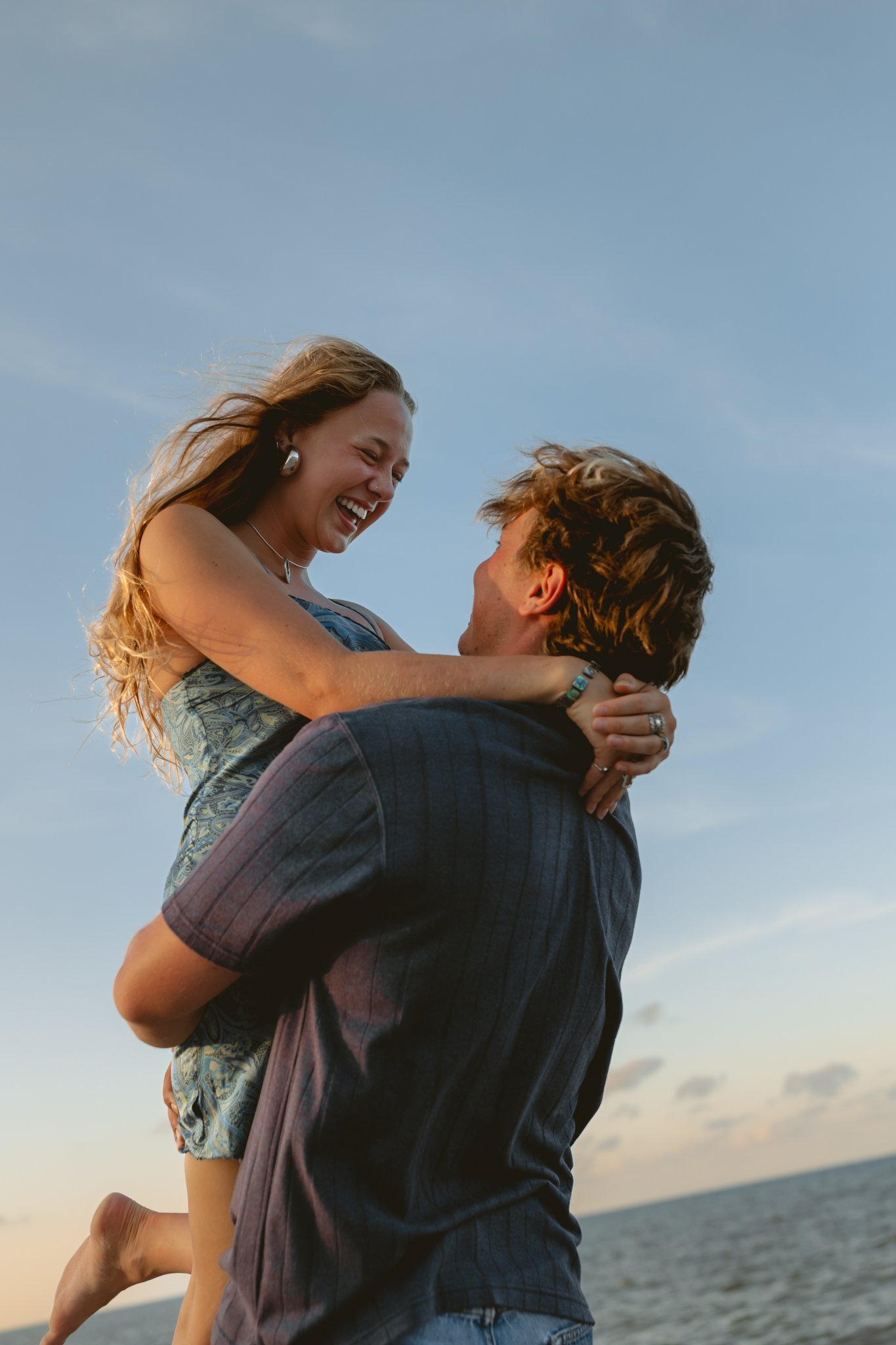 A couple at the beach, the man lifting the woman, both smiling and enjoying sunset, with ocean and sky in background.