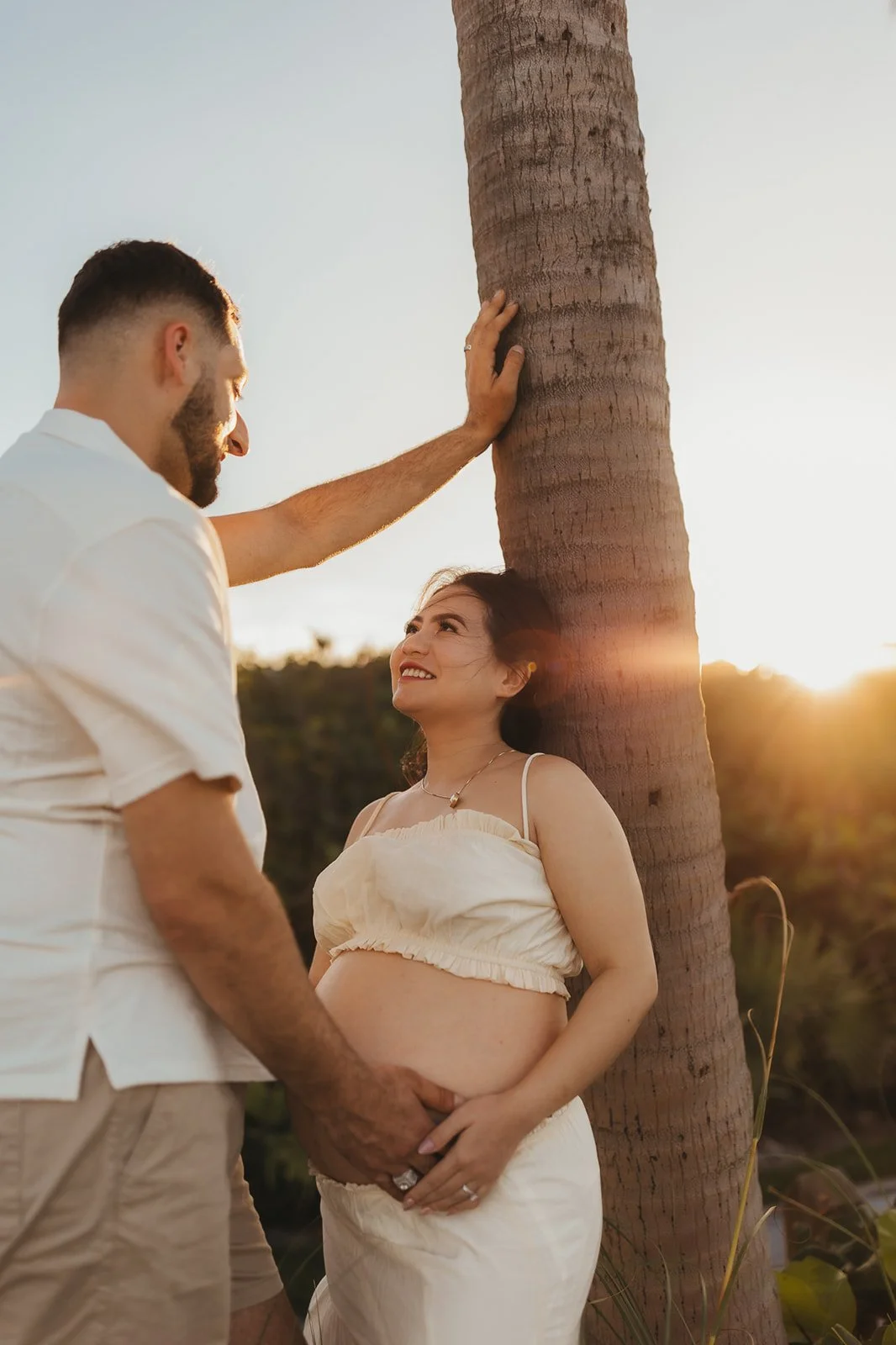 A pregnant woman leaning against a palm tree, smiling at her partner who is touching the tree with one hand during sunset.
