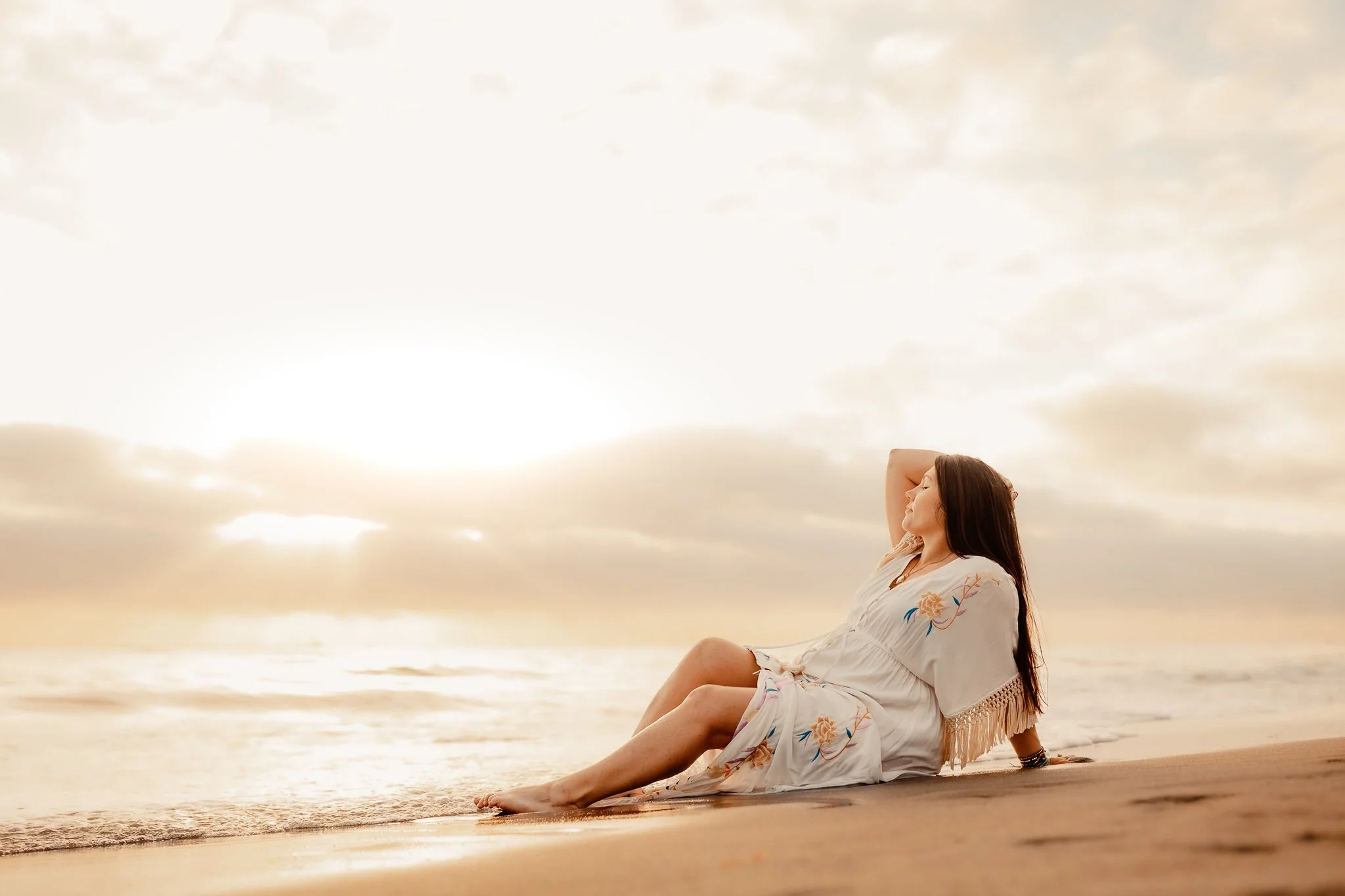 Woman relaxing on sandy beach at sunrise or sunset with her eyes closed and hand behind her head, wearing a white dress with floral embroidery.