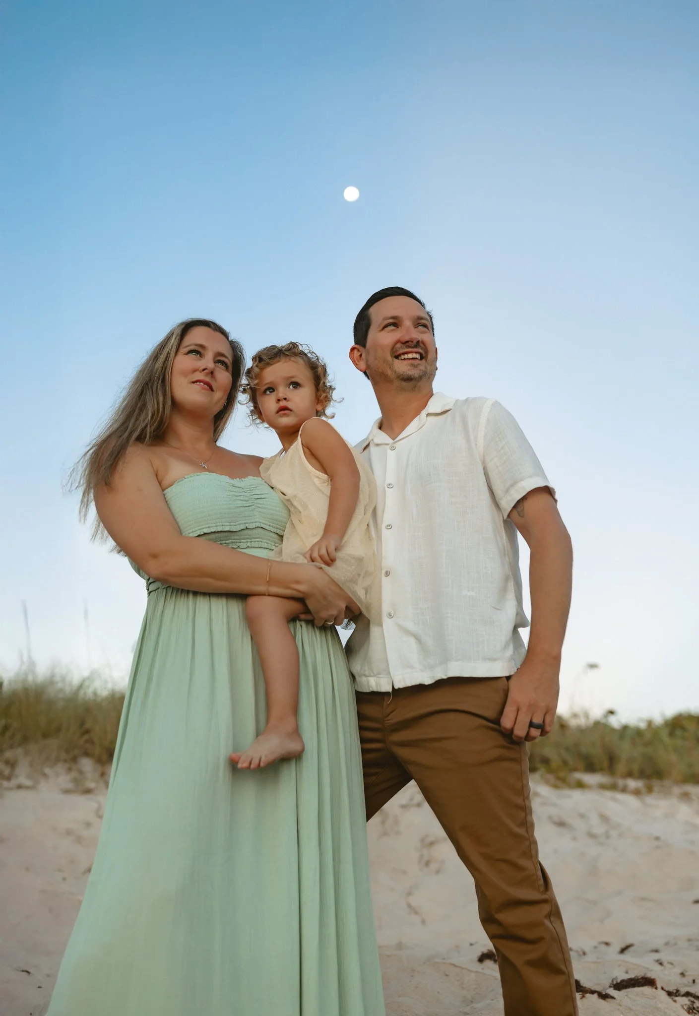 A family of three, a woman, a man, and a young girl, standing on a beach during twilight with the moon visible in the sky.
