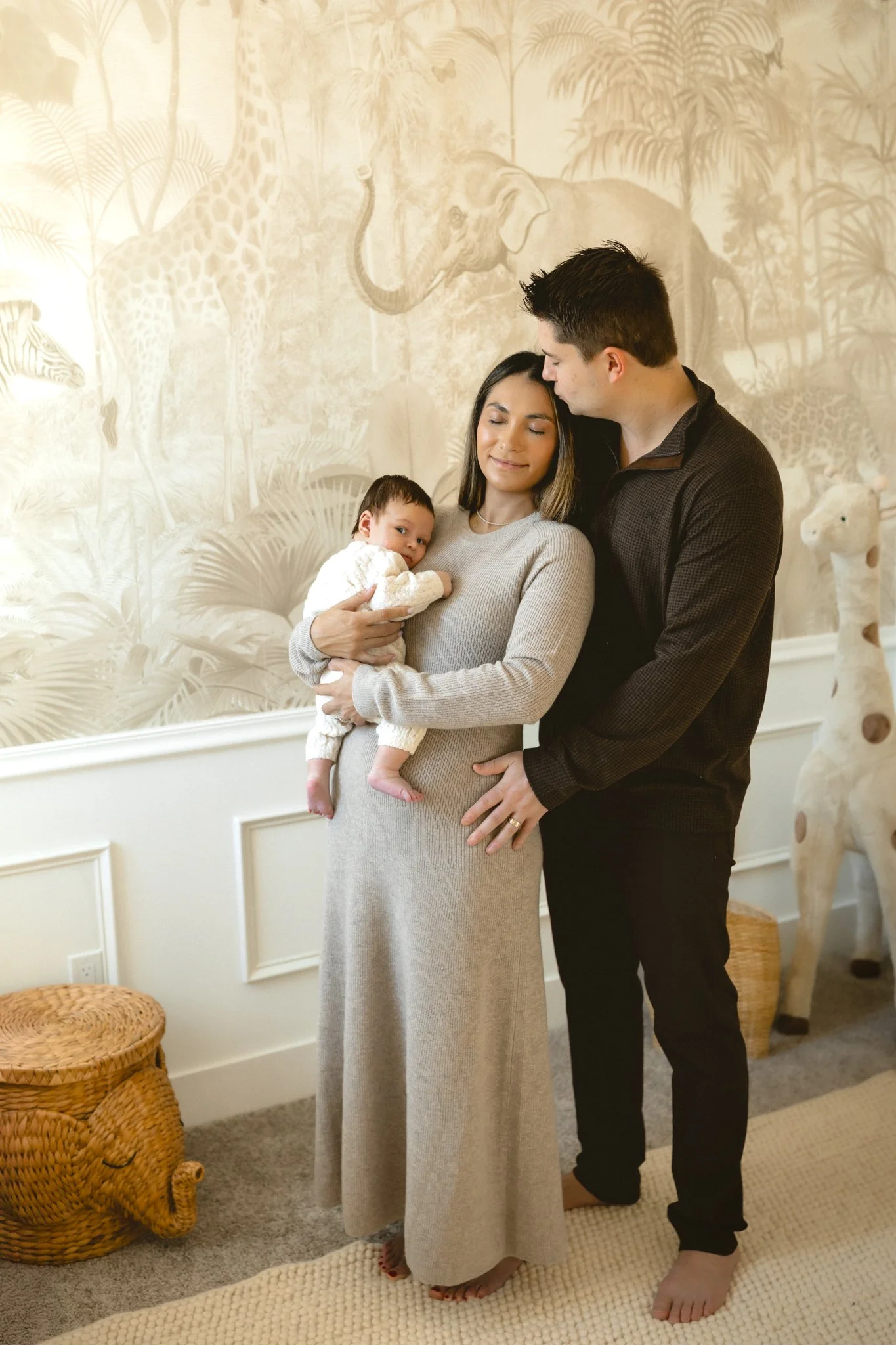 A family of three standing together indoors in front of a jungle-themed wall with animal illustrations, including giraffes and an elephant. The woman is holding a young baby, and the man is standing close, touching her waist.