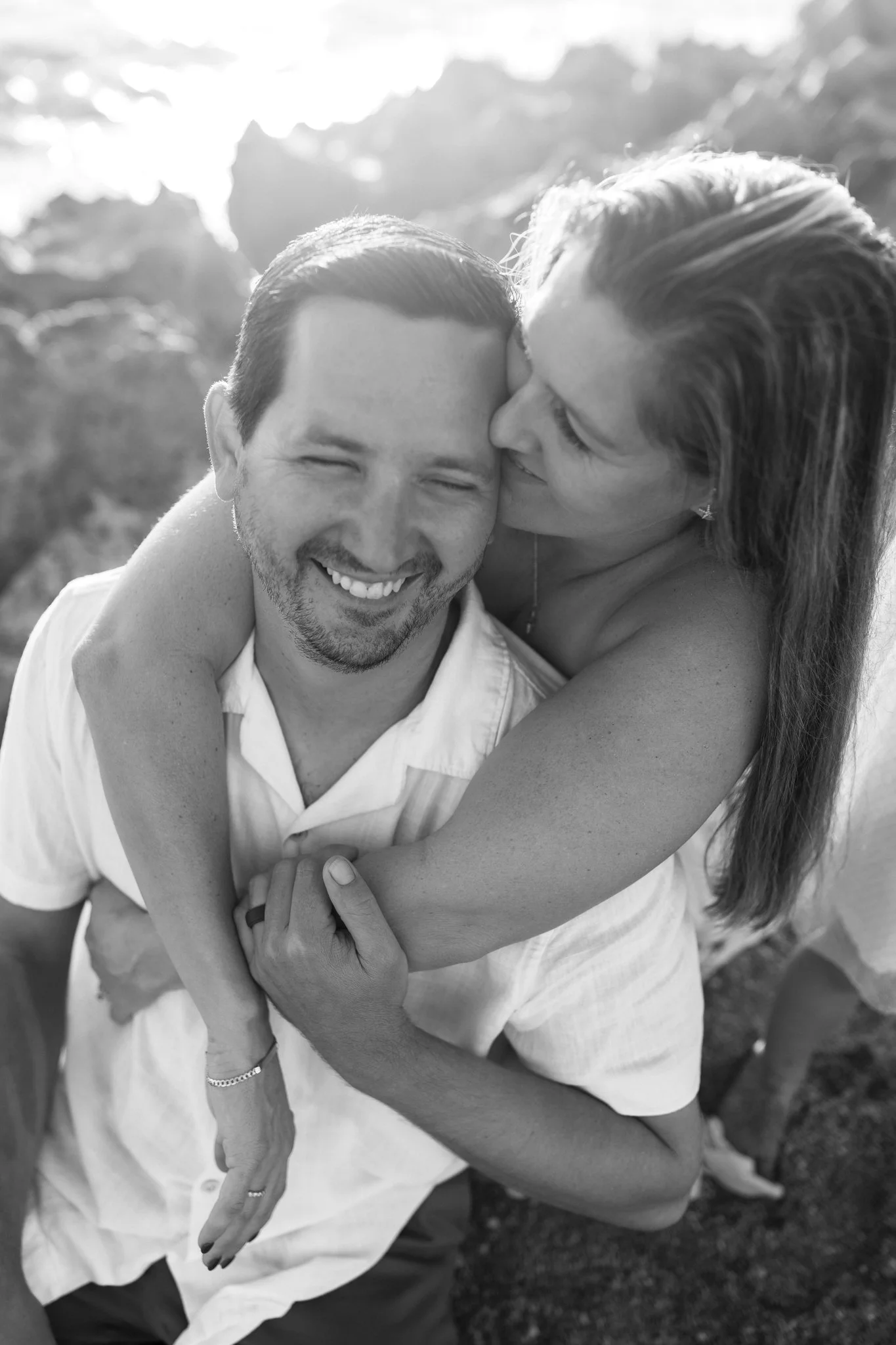 A black and white photo of a happy couple, with the woman kissing the man's cheek. The man is smiling with eyes closed, and the woman has her arms around his shoulders. They are outdoors with natural scenery in the background.