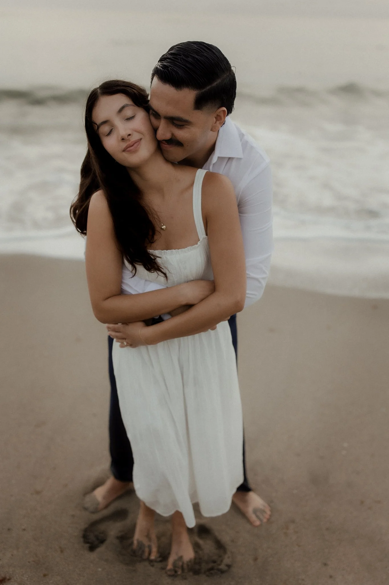 A couple hugging on the beach, with the woman in a white dress and the man in a white shirt and dark pants, standing barefoot in the sand.