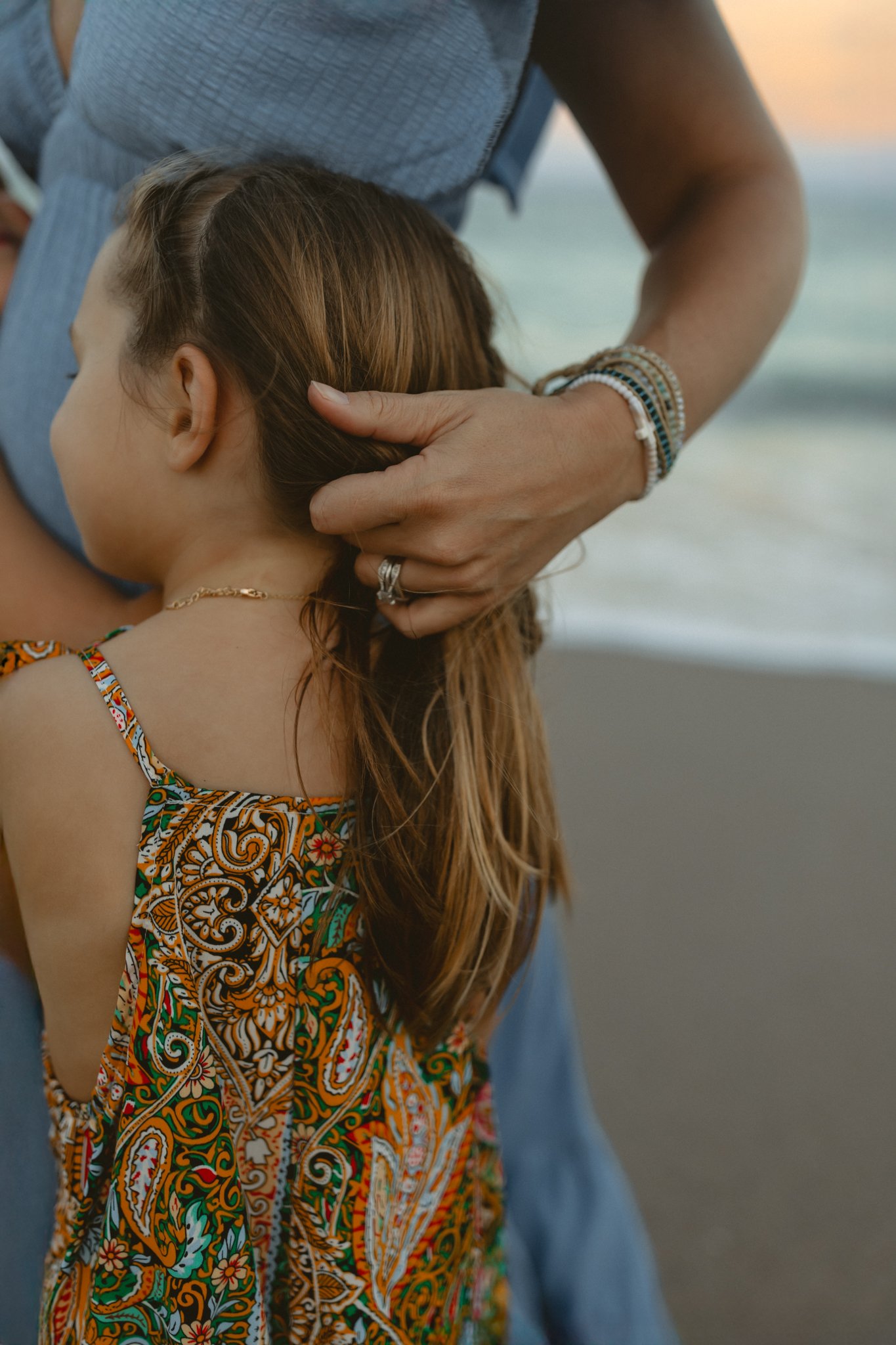 A woman with brown hair wearing a colorful patterned dress is holding a young girl with light brown hair and fair skin. The woman is wearing multiple bracelets and rings, and the girl is wearing a gold necklace. The background shows a beach with the 