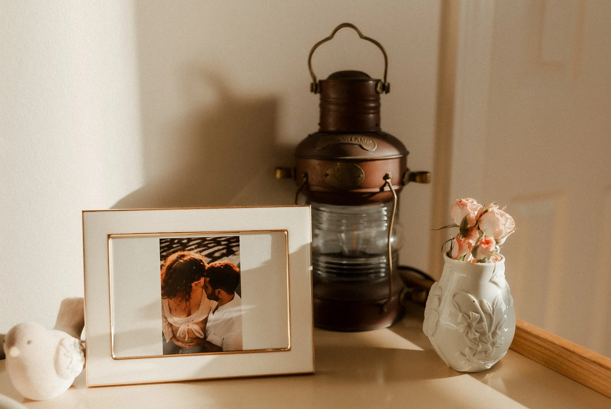A framed photograph of a couple leaning in close, a vintage lantern, a white vase with pink flowers, and a small ceramic bird on a wooden surface.