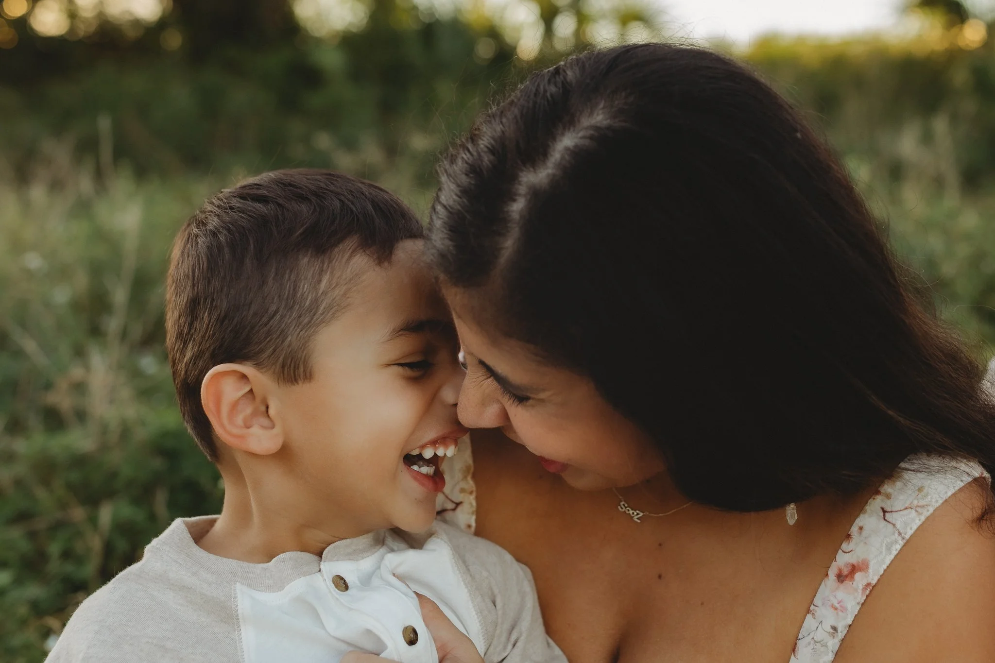 A woman and a young boy are closely face to face, smiling and sharing a joyful moment outdoors.