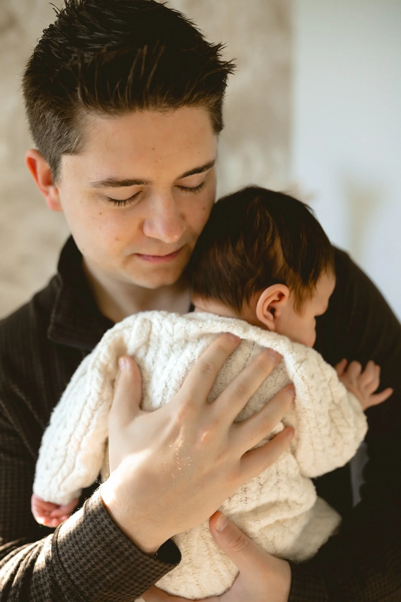 A young man holding a newborn baby close, both with their eyes closed, showing affection and tenderness.