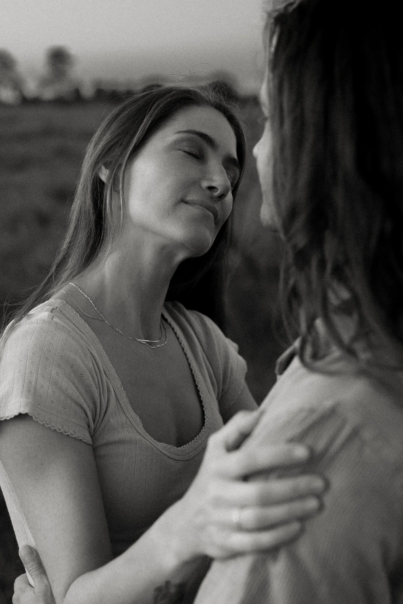 A black and white photo of two women close together, with one woman gently resting her face against the other's. They appear to be affectionate, with the woman on the left smiling softly and her eyes closed. The background shows an outdoor setting wi