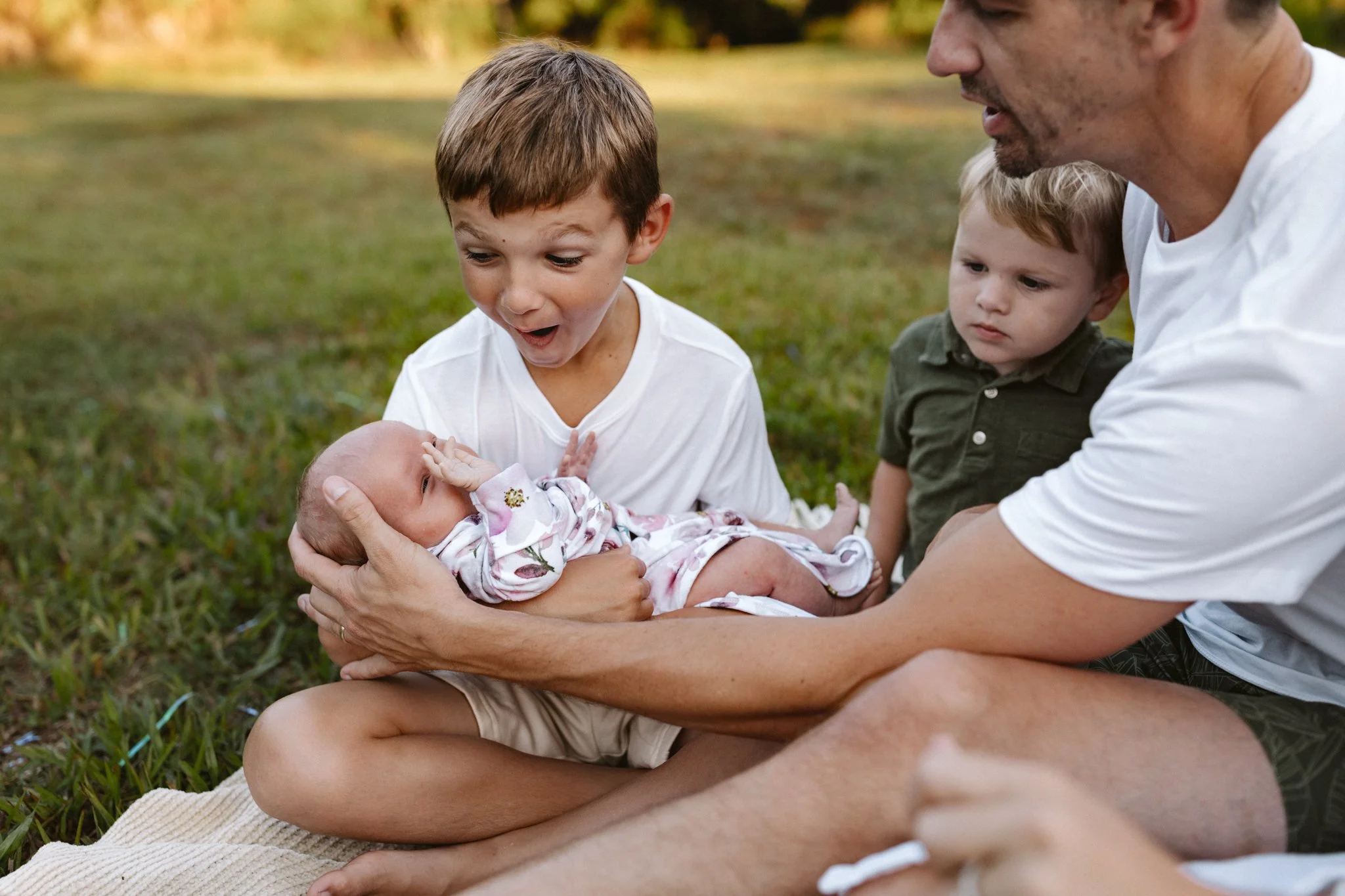 A man and three children sitting on grass outdoors, with a baby being cradled by the man, all looking at the baby with surprise and curiosity.
