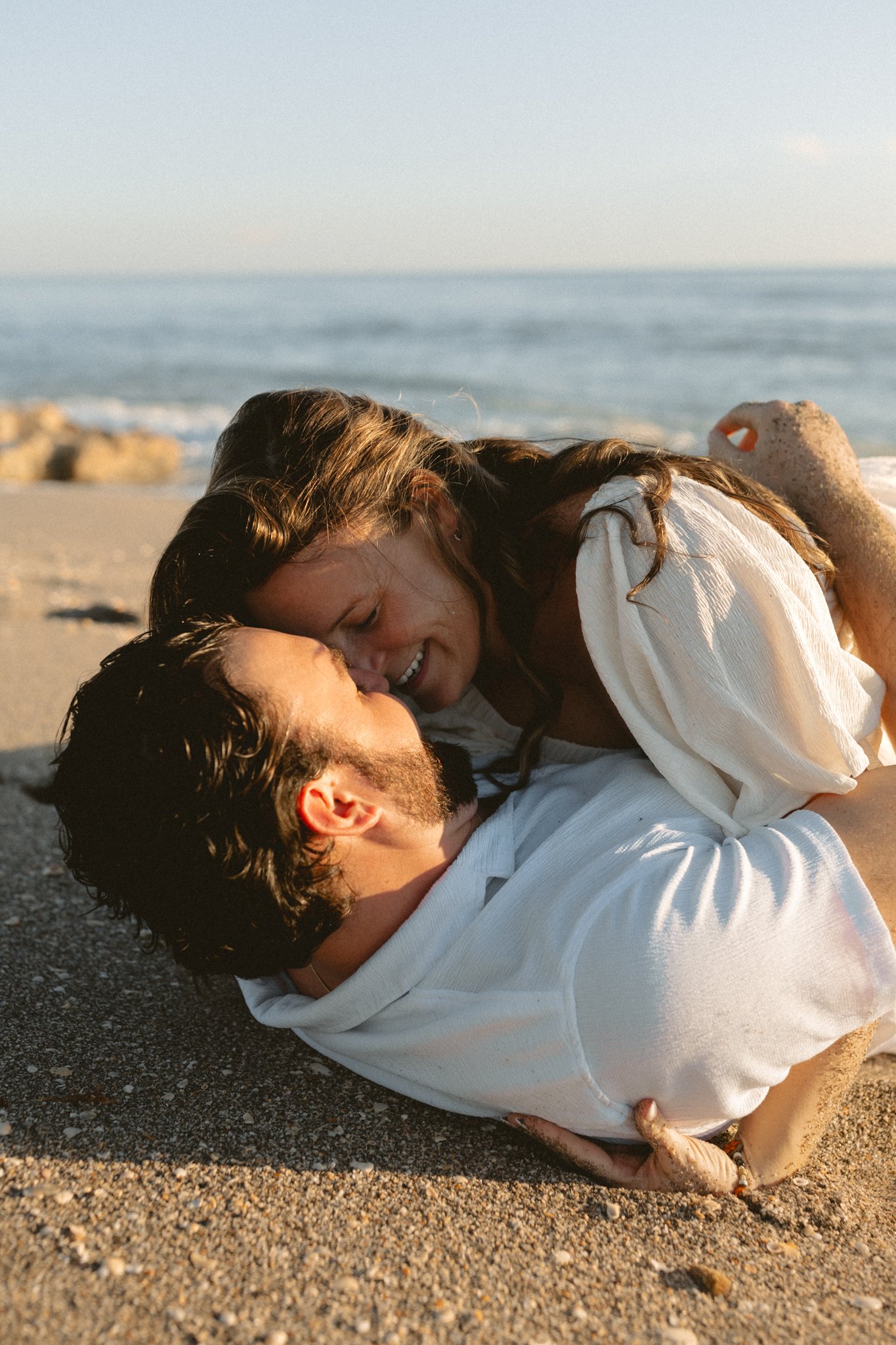 A couple lying on a sandy beach, smiling and touching noses, with the ocean and rocks in the background.