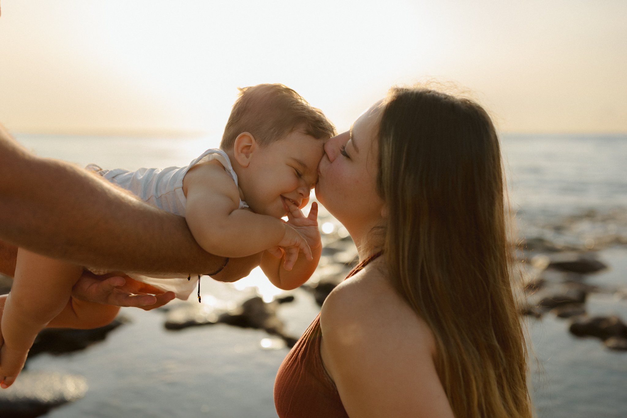 A woman holding a smiling baby close to her face on a beach at sunset.