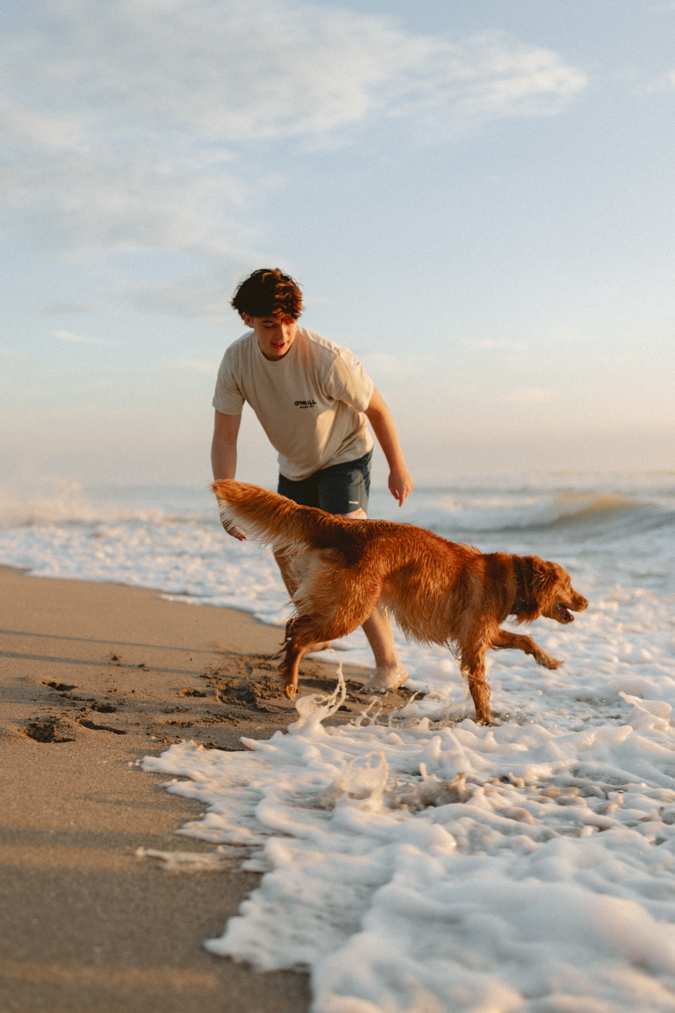 A young man and a golden retriever dog playing and running along the shoreline at the beach with waves crashing around them during sunset.
