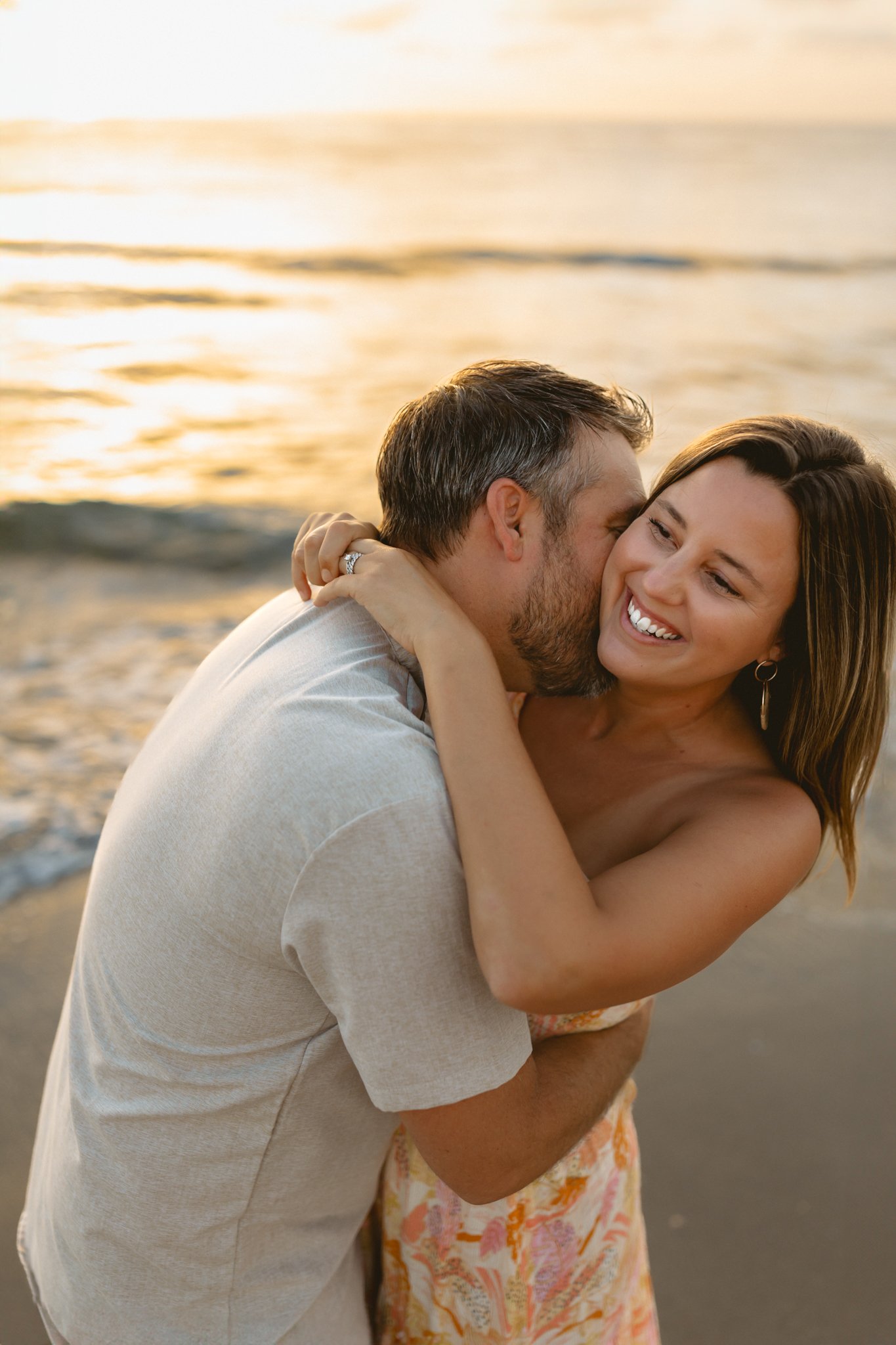 A smiling woman with shoulder-length brown hair and earrings hugging a man with short gray hair and beard on a beach at sunset, with waves in the background.