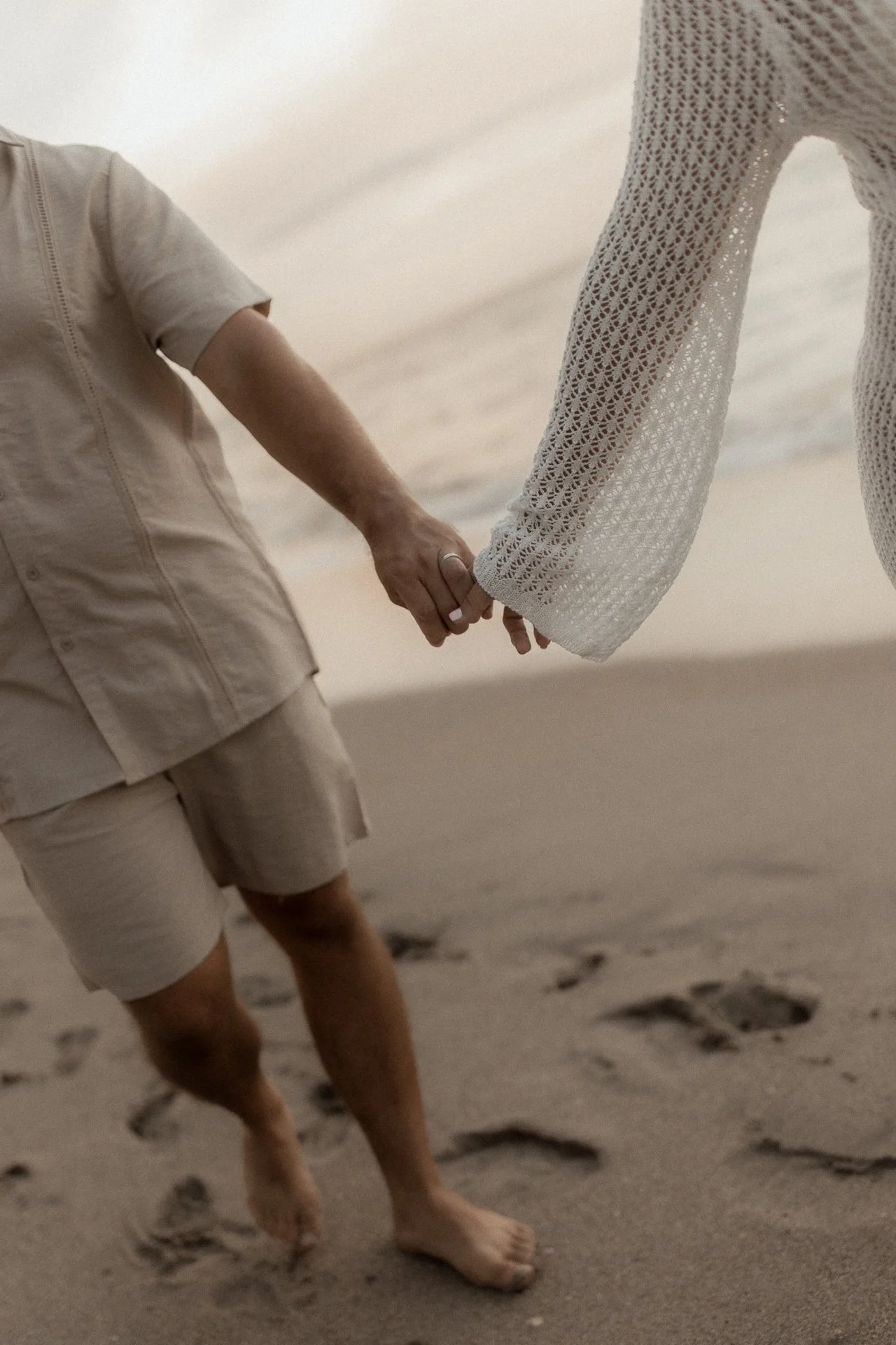 Two people holding hands on the beach, with one person wearing a light-colored shirt and shorts, the other in a white, crochet-style top. Bare feet are visible, and footprints are in the sand.