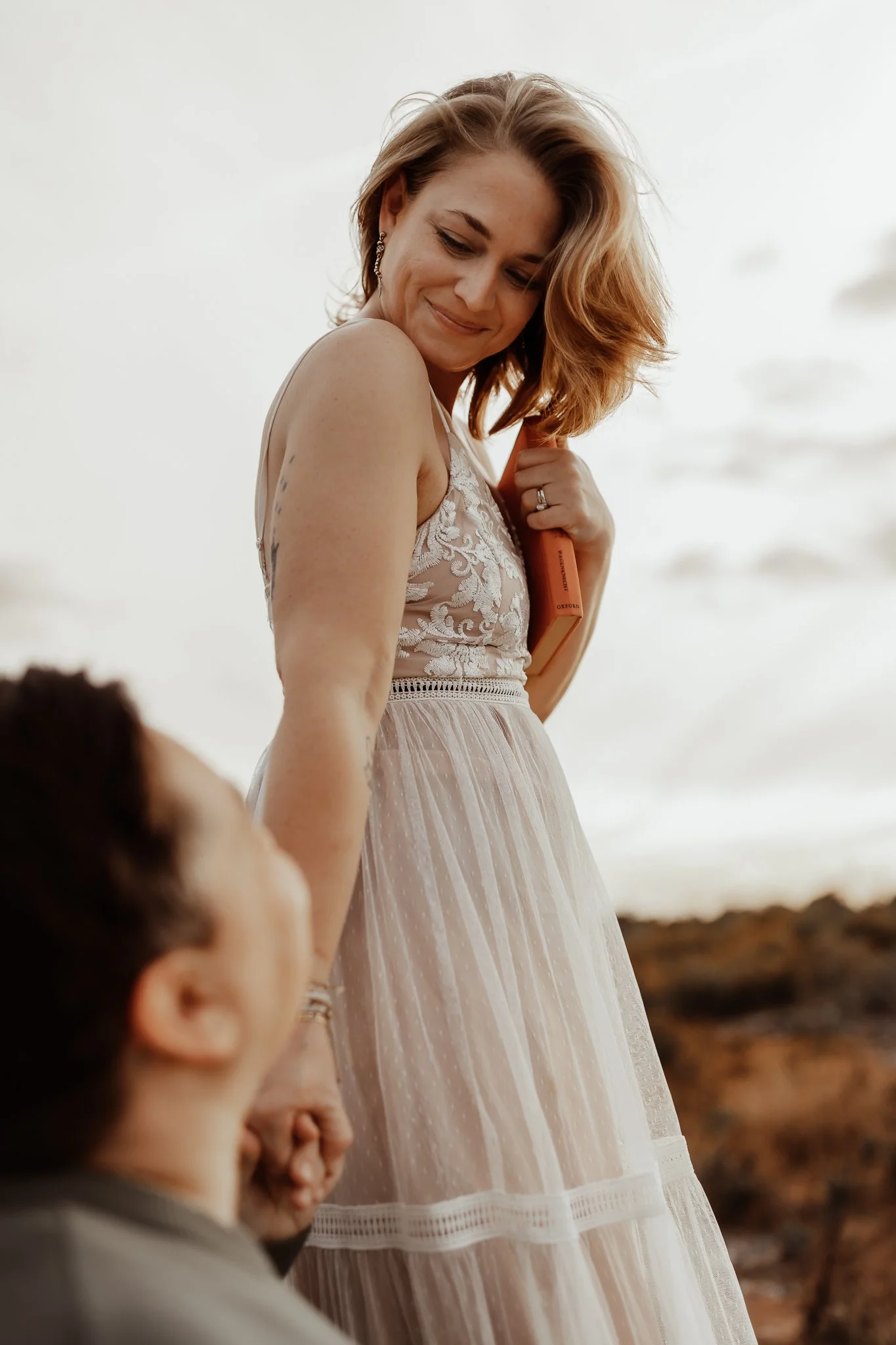 A woman in a white lace dress standing on a beach holding a person's hand. She is smiling and looking down, with a book tucked under her arm. The scene is set against a cloudy sky and rocky shoreline.