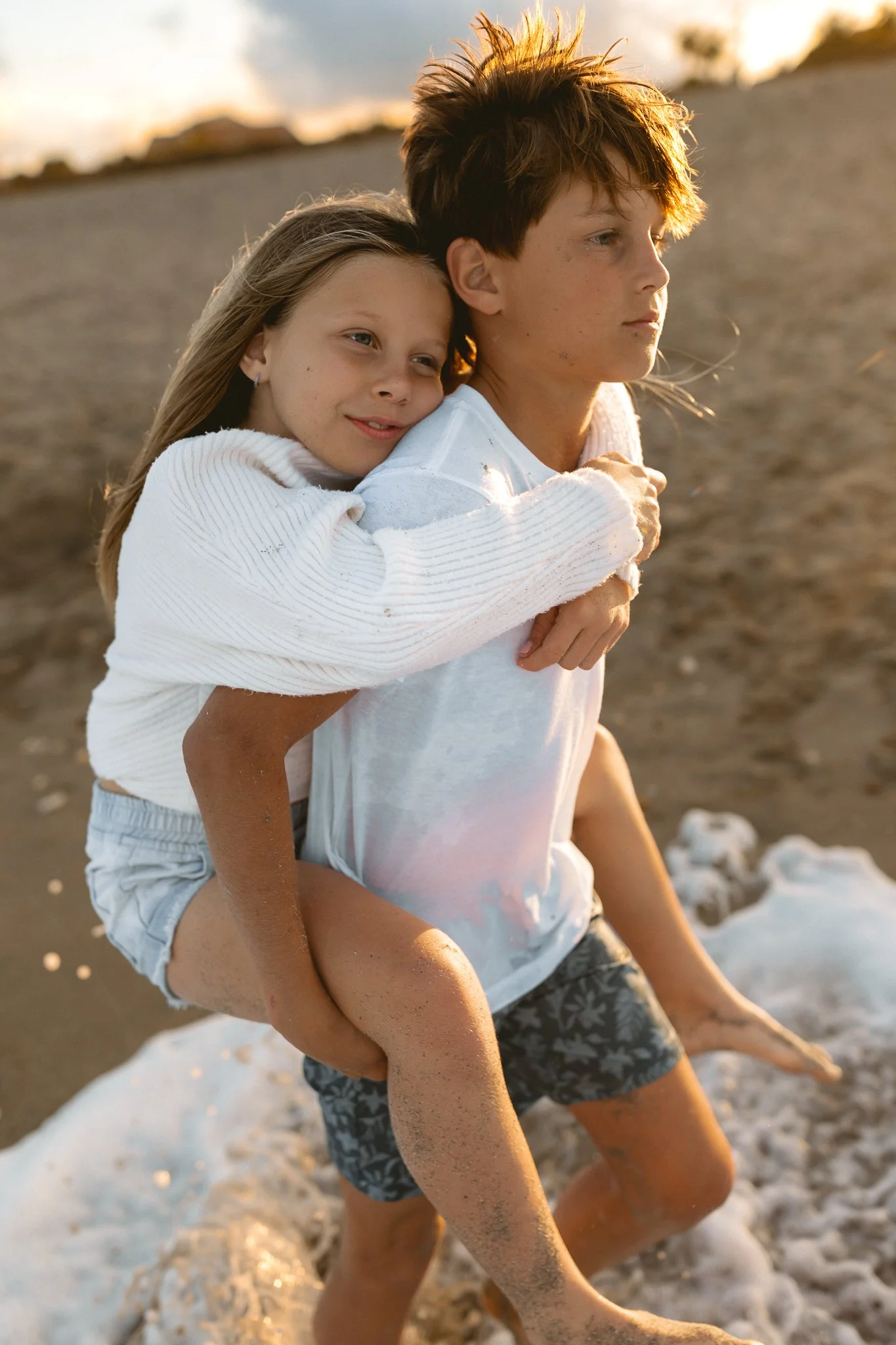 a young boy carrying his young sister on his back while walking on the beach