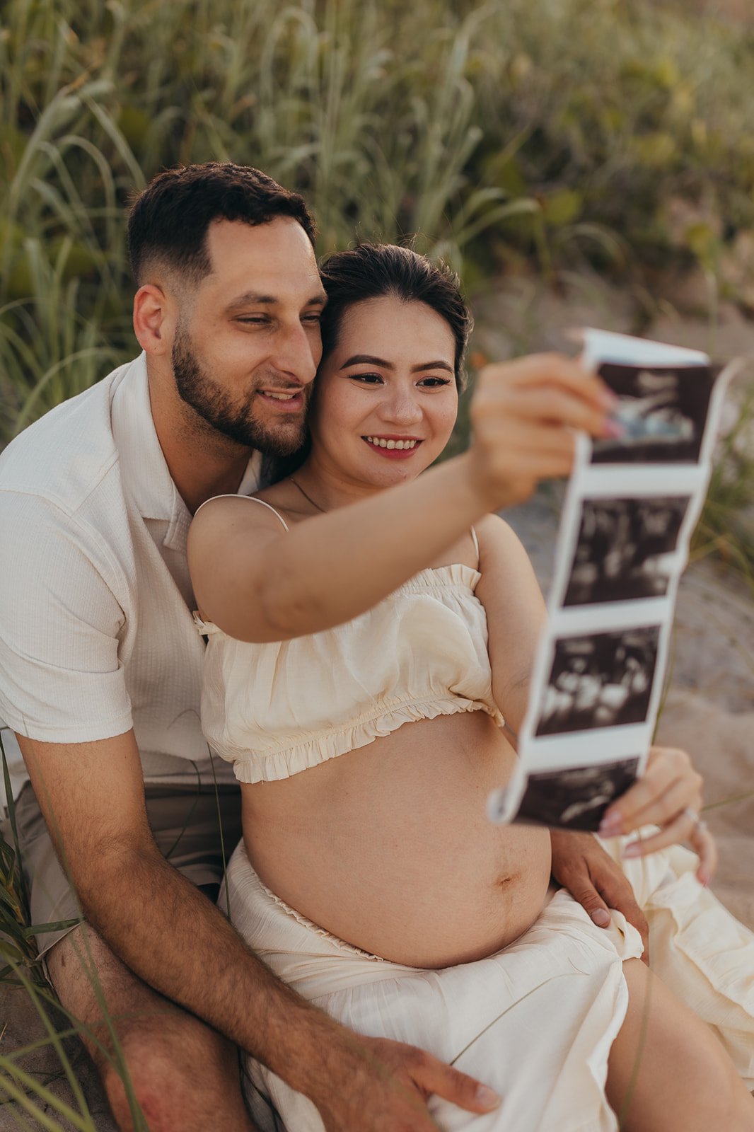 A happy pregnant woman and her partner taking a selfie together on a beach, sitting among tall grass and sand.