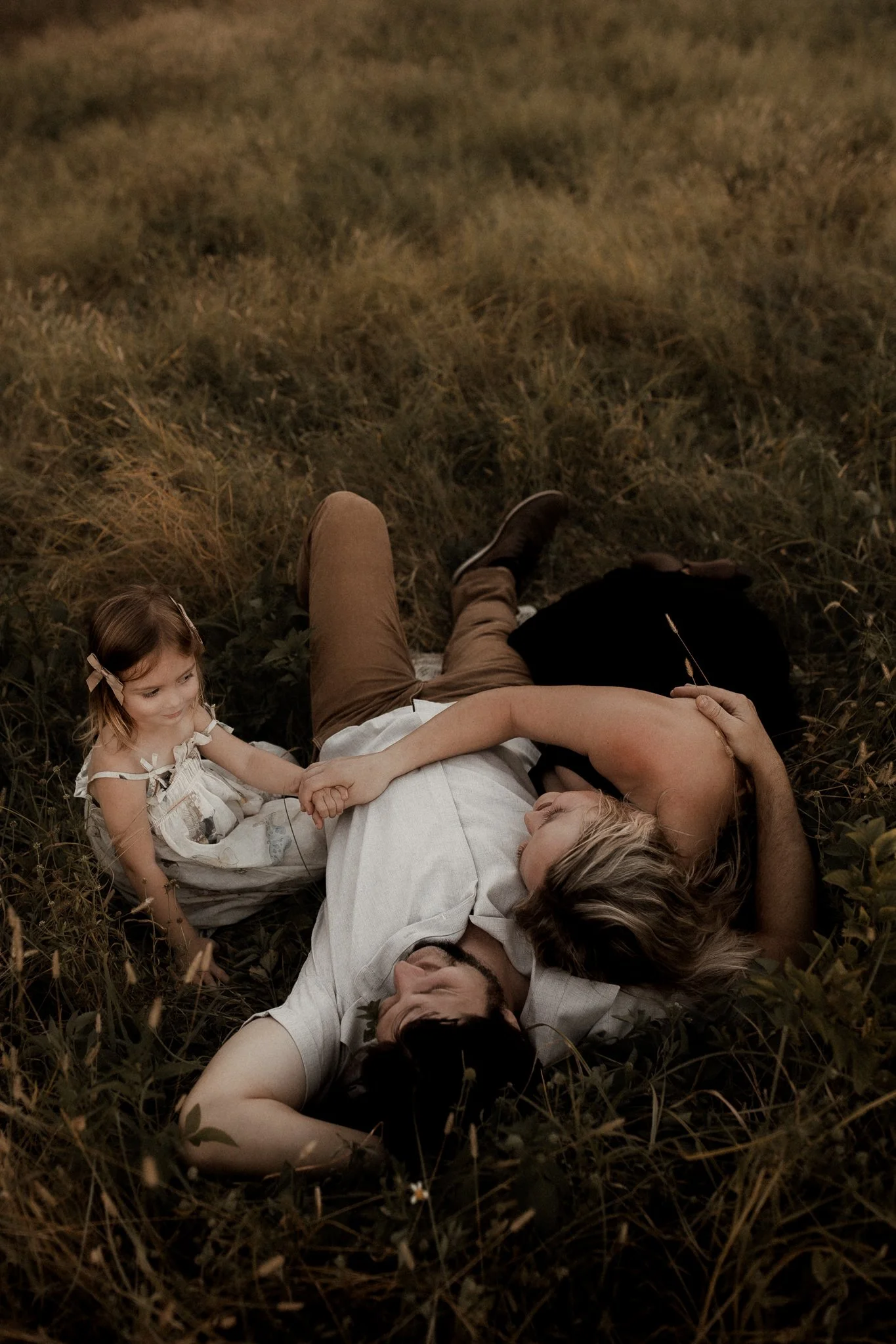 A family lying on the grass in a field. A woman and man are lying on their sides, holding hands. A young girl, sitting up, is holding the woman's hand and smiling.
