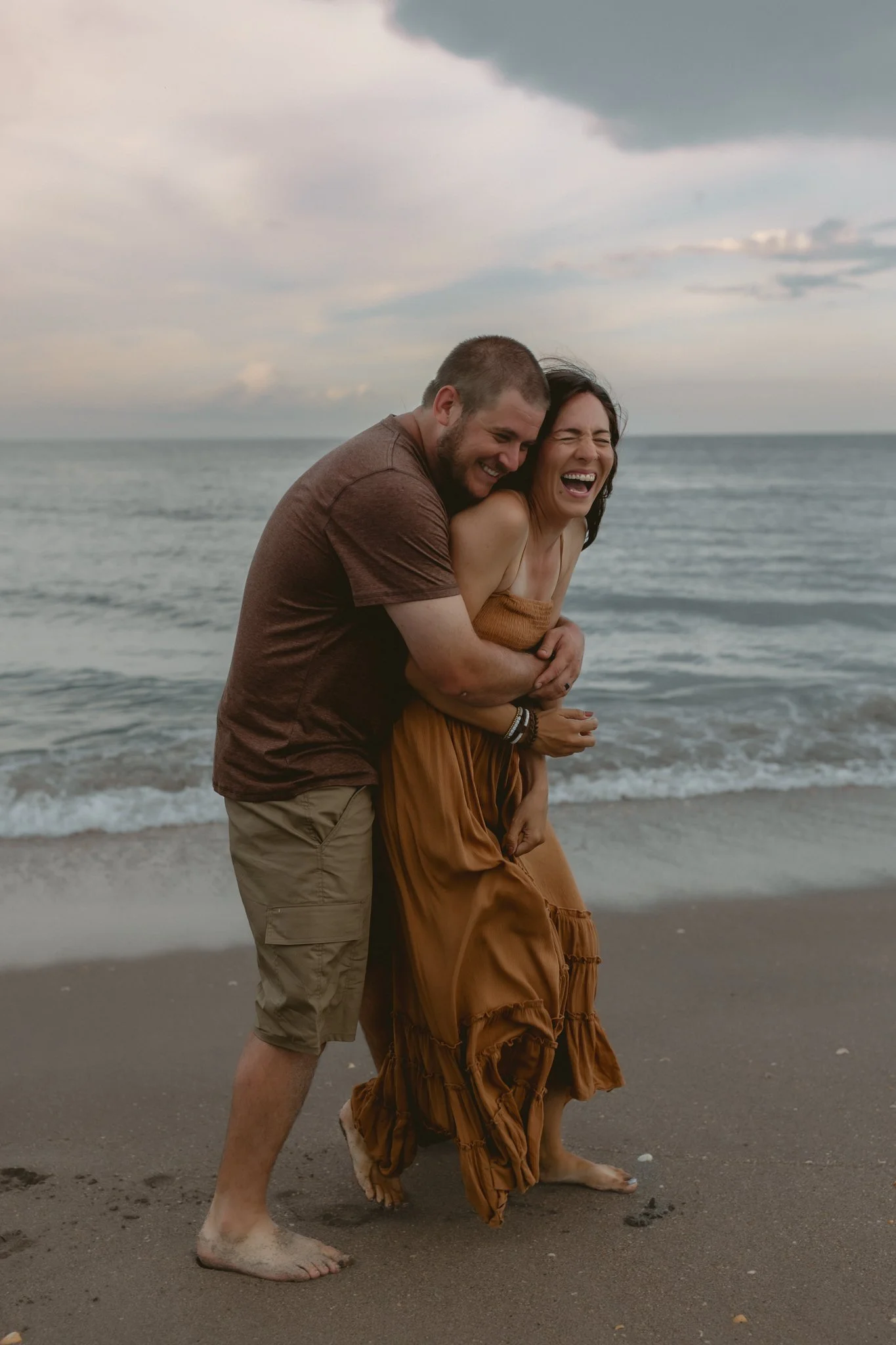 A joyful couple laughing and embracing on a beach with ocean waves and a cloudy sky in the background.