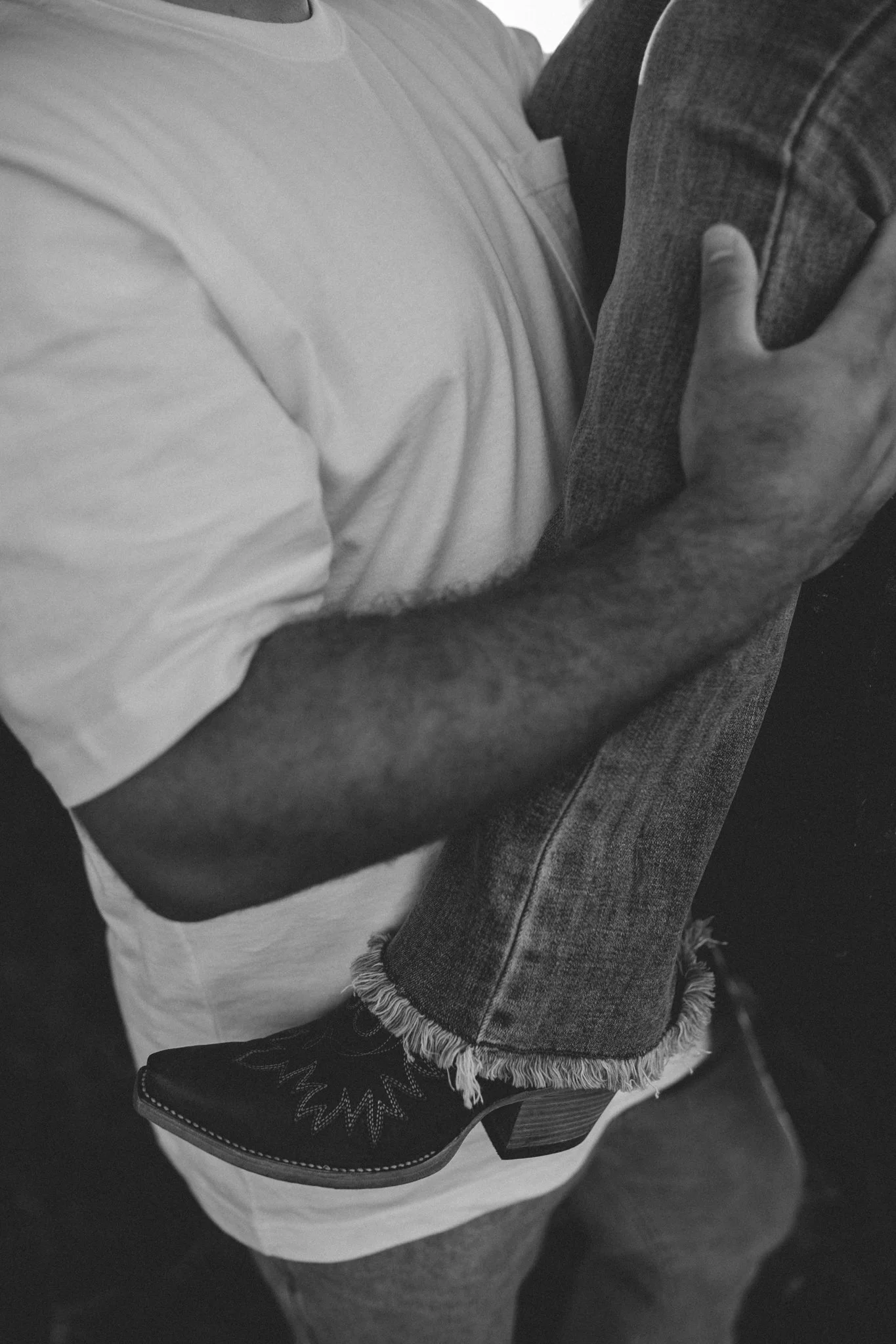 A close-up black and white photo of a person wearing a white t-shirt and jeans with a woman on their hip, resting her leg on the person's waist. The woman's shoe features embroidered designs, and her hand rests on the person's shoulder.